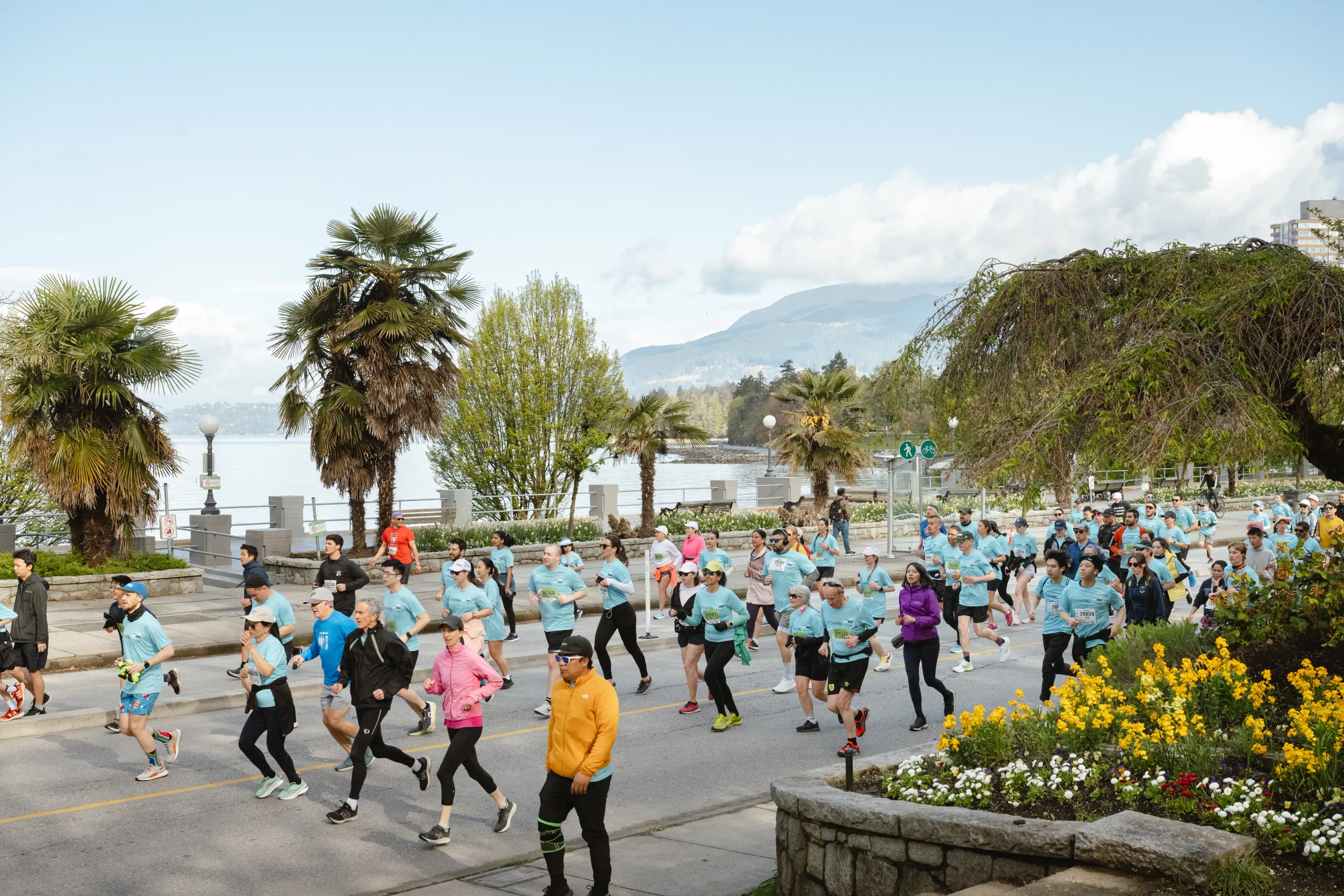 A large group of people running outdoors on a road near palm trees and a waterfront with mountains in the background.