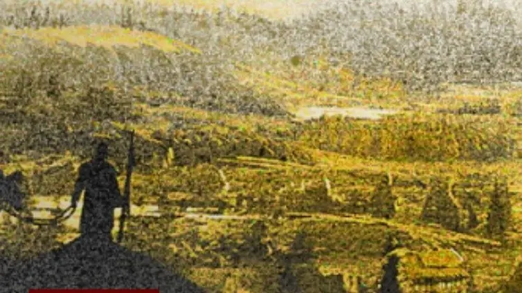 Silhouette of a person holding a rifle overlooking a vast, yellow-tinted landscape with hills and scattered buildings.