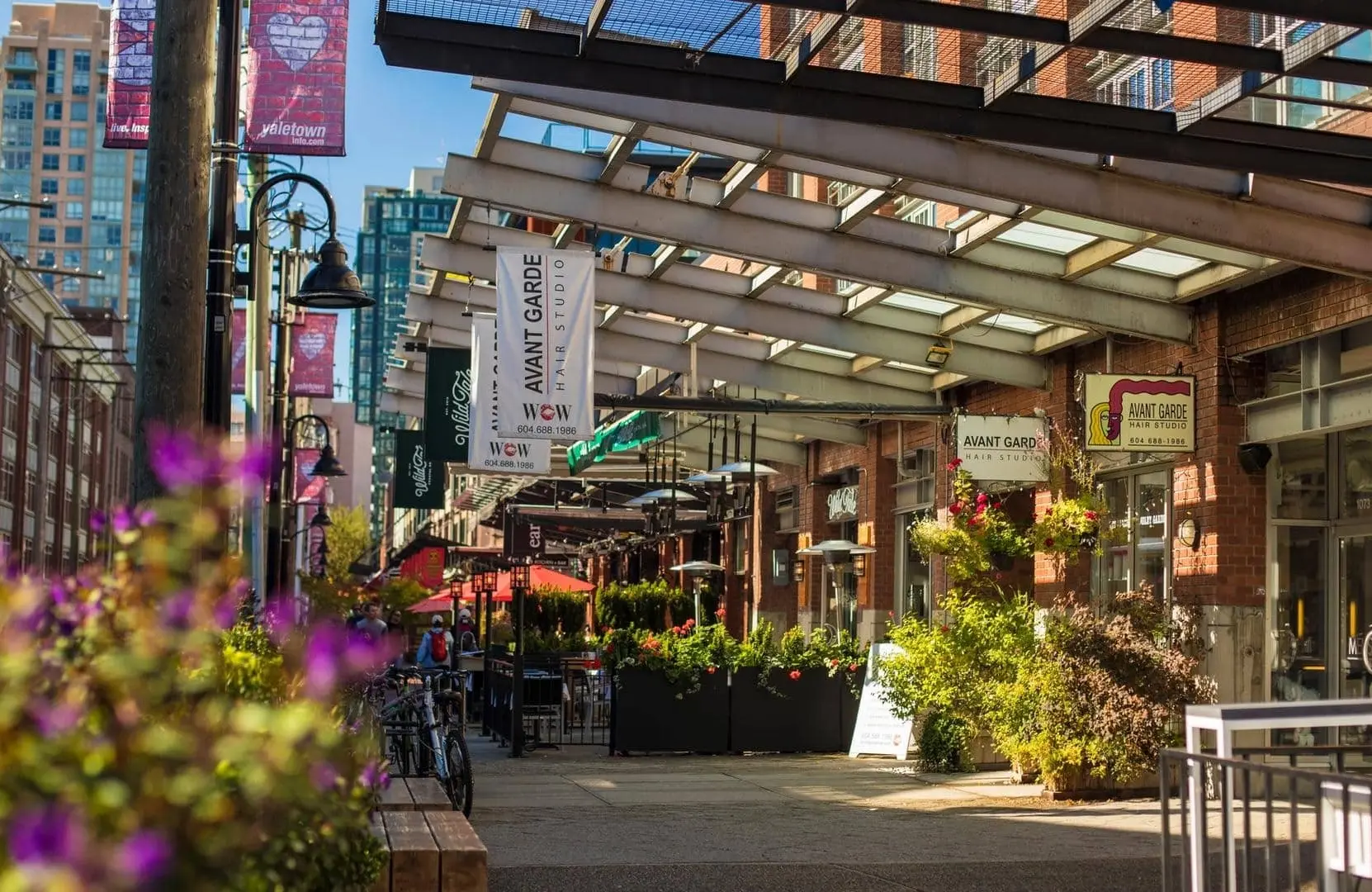 A bright Vancouver sidewalk with storefronts, plants, pink flowers, and patio spots