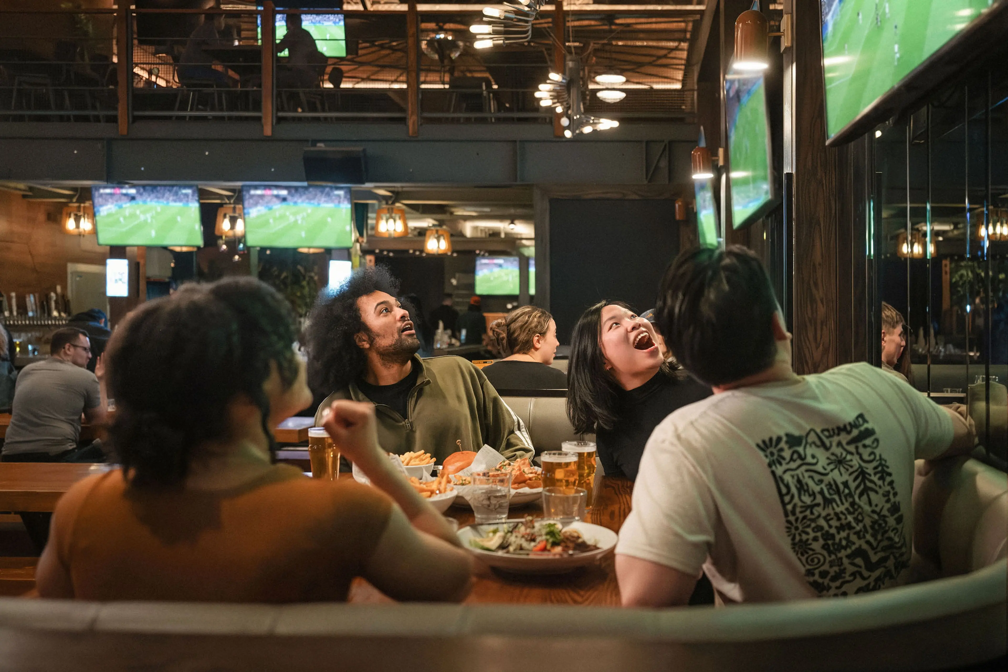 Four people sitting at a restaurant booth watching multiple TVs showing a soccer game, with food and drinks on the table.