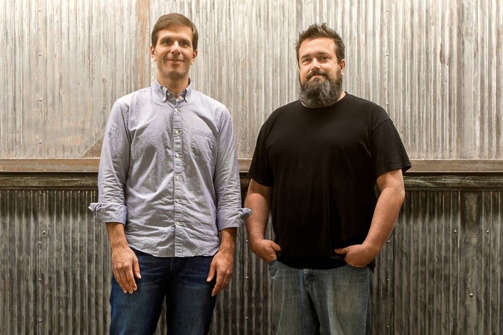 Two people standing in front of a corrugated metal wall, one wearing a light button-up shirt and jeans, the other wearing a black t-shirt and jeans with hands in pockets.