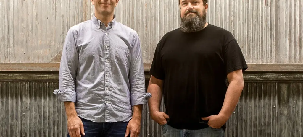Two people standing in front of a corrugated metal wall, one wearing a light button-up shirt and jeans, the other wearing a black t-shirt and jeans with hands in pockets.