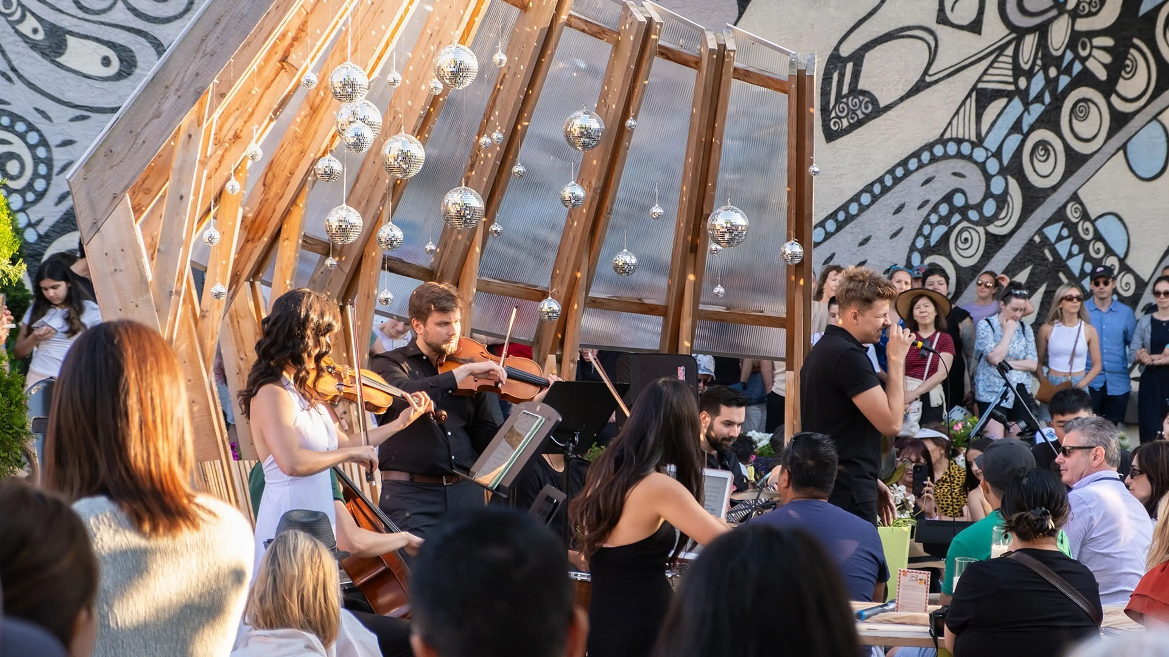 Musicians playing string instruments under a wooden structure with hanging disco balls, surrounded by a seated audience outdoors.