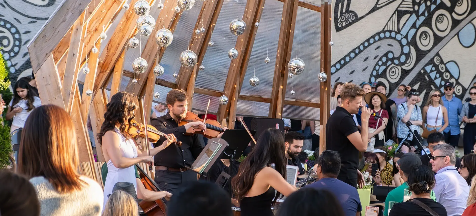 Musicians playing string instruments under a wooden structure with hanging disco balls, surrounded by a seated audience outdoors.
