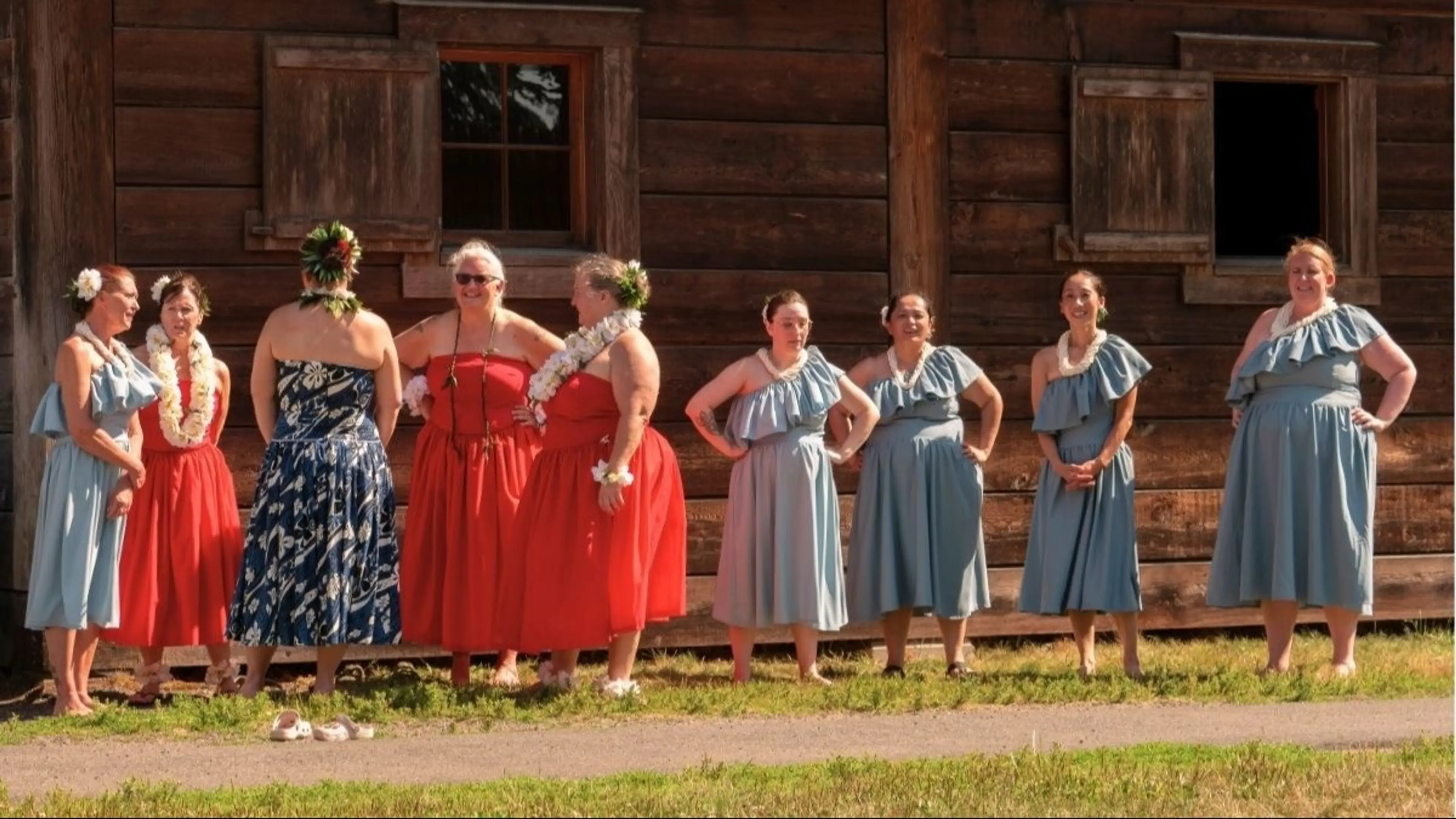 Ten people wearing red and blue dresses with floral leis, standing outside a wooden building on grass.