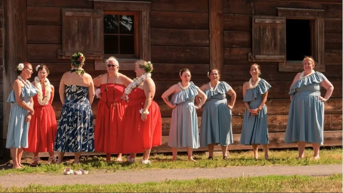 Ten people wearing red and blue dresses with floral leis, standing outside a wooden building on grass.