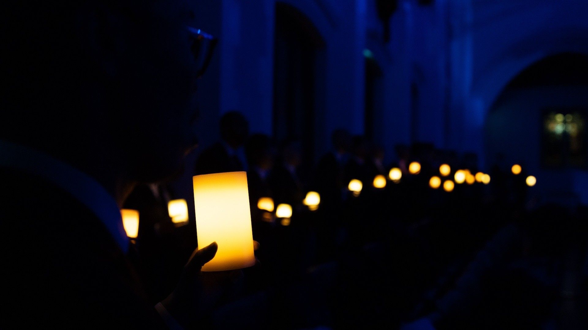 A person holding a glowing candle in a dark room with many other glowing candles in the background.