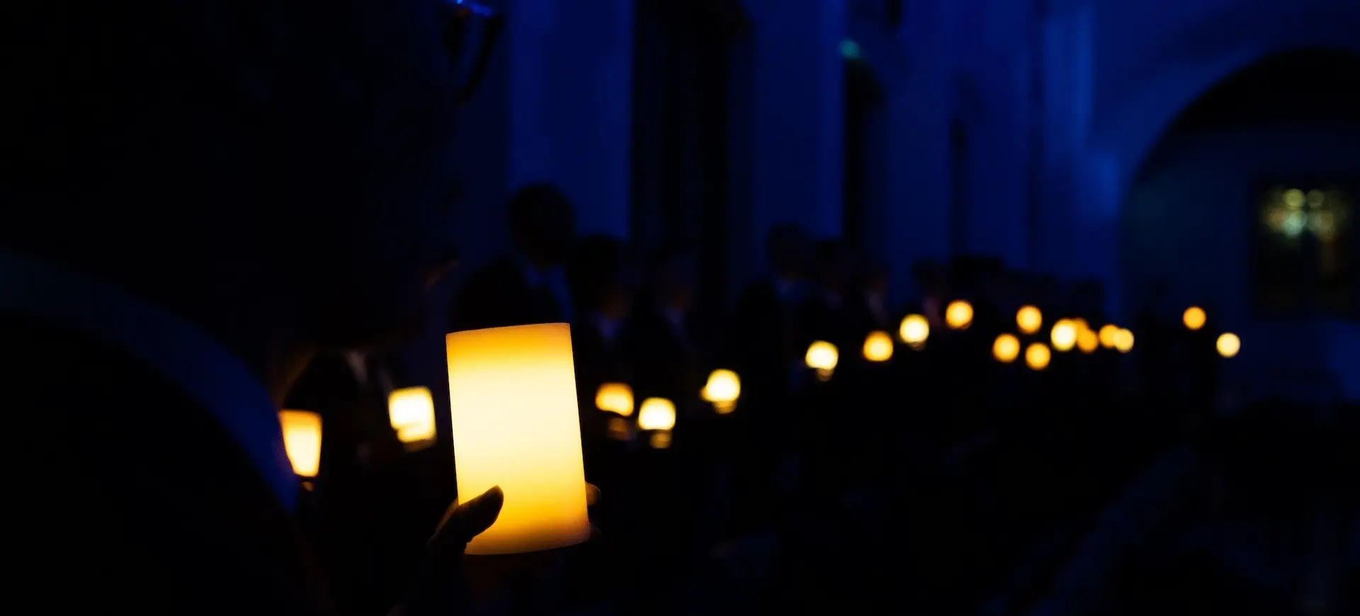 A person holding a glowing candle in a dark room with many other glowing candles in the background.