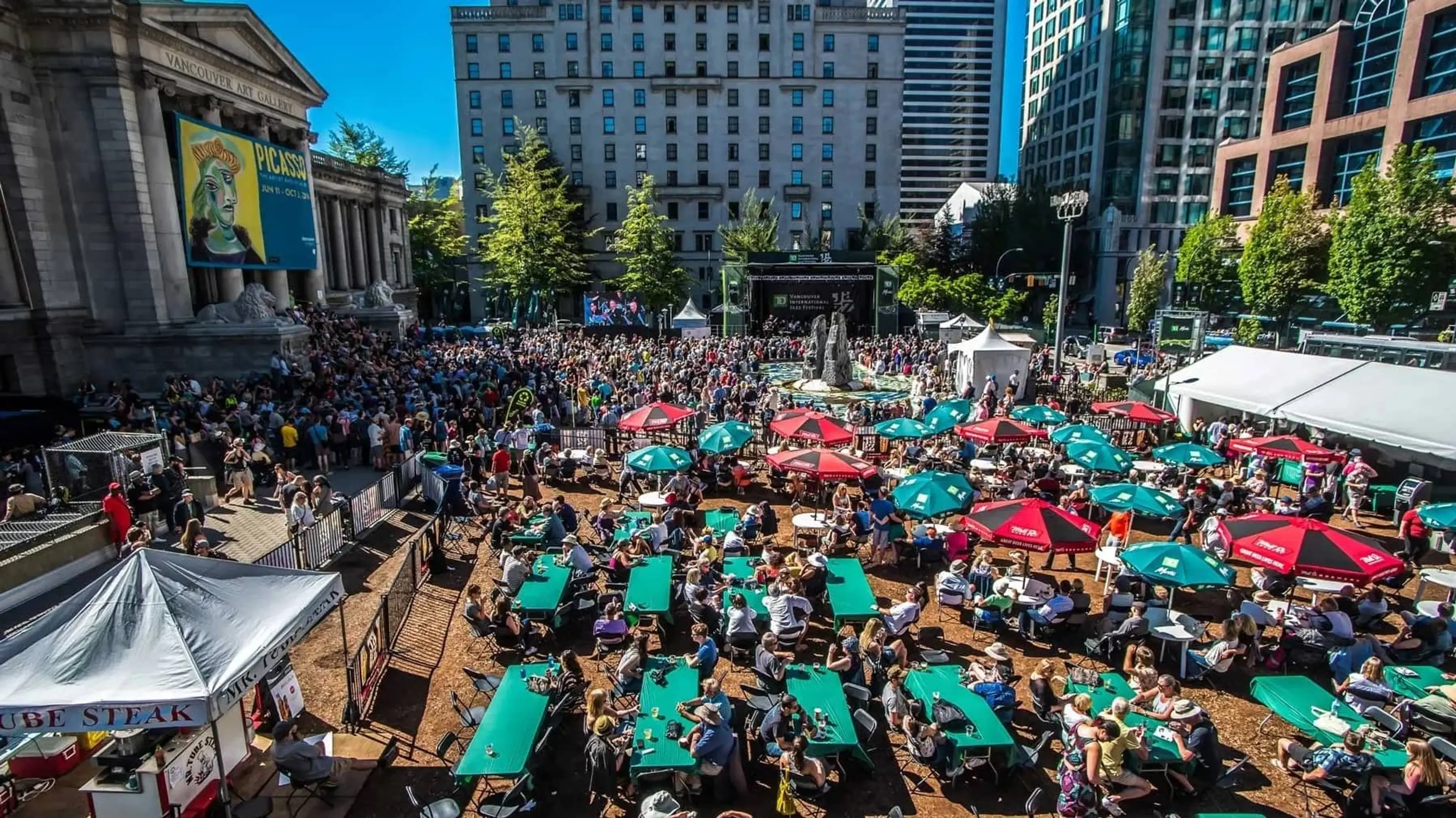 A crowd of people stand in front of an outdoor urban stage with umbrellas, tables, and tents nearby.
