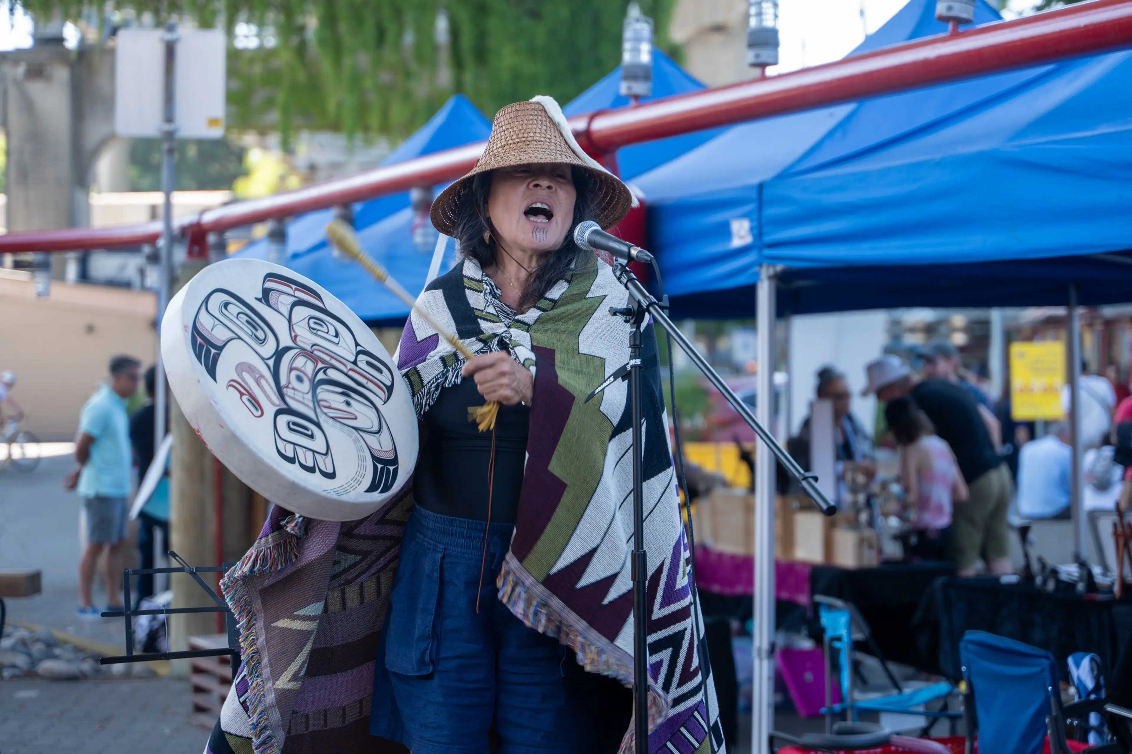 A person wearing a woven hat and patterned shawl sings into a microphone while holding a decorated drum and drumstick at an outdoor event with blue tents.