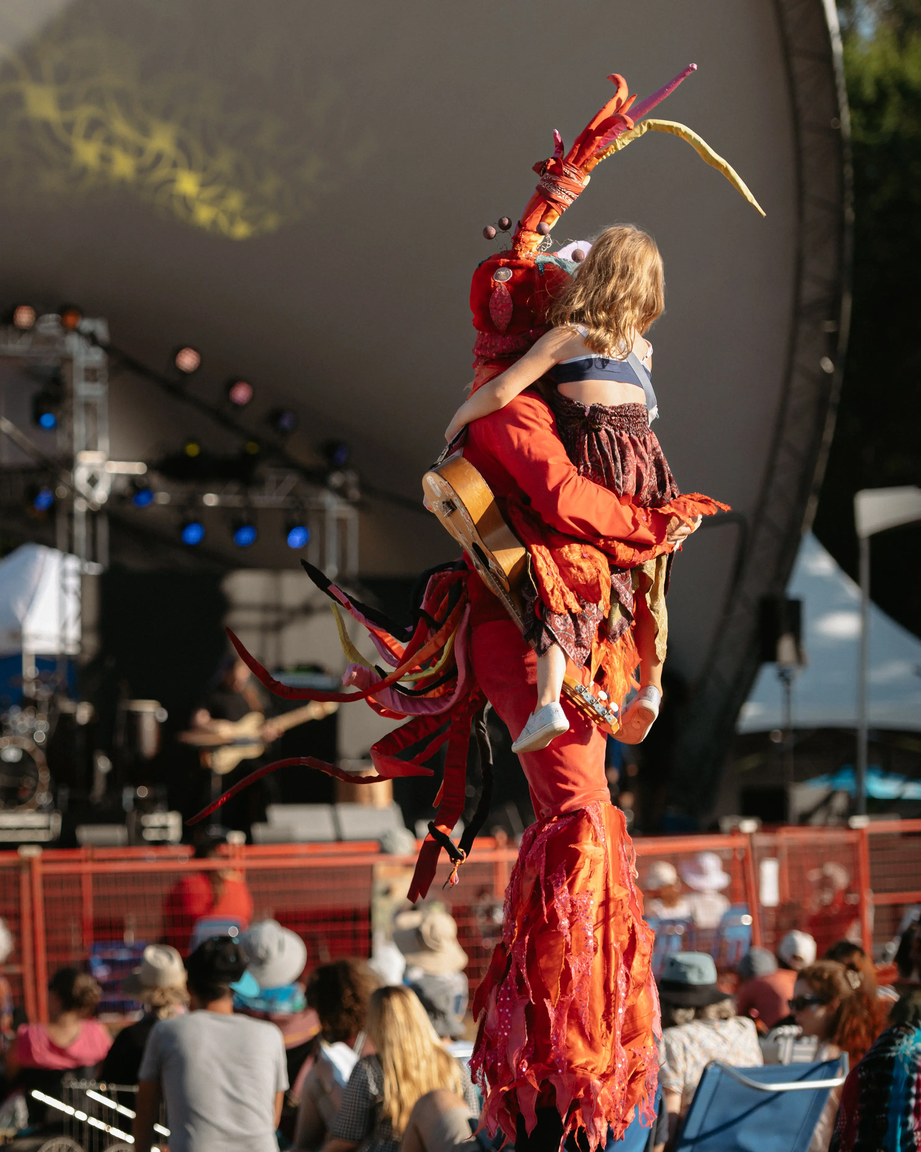 Person in a red lobster costume holding a child in a dress at an outdoor event with seated audience and stage in the background.