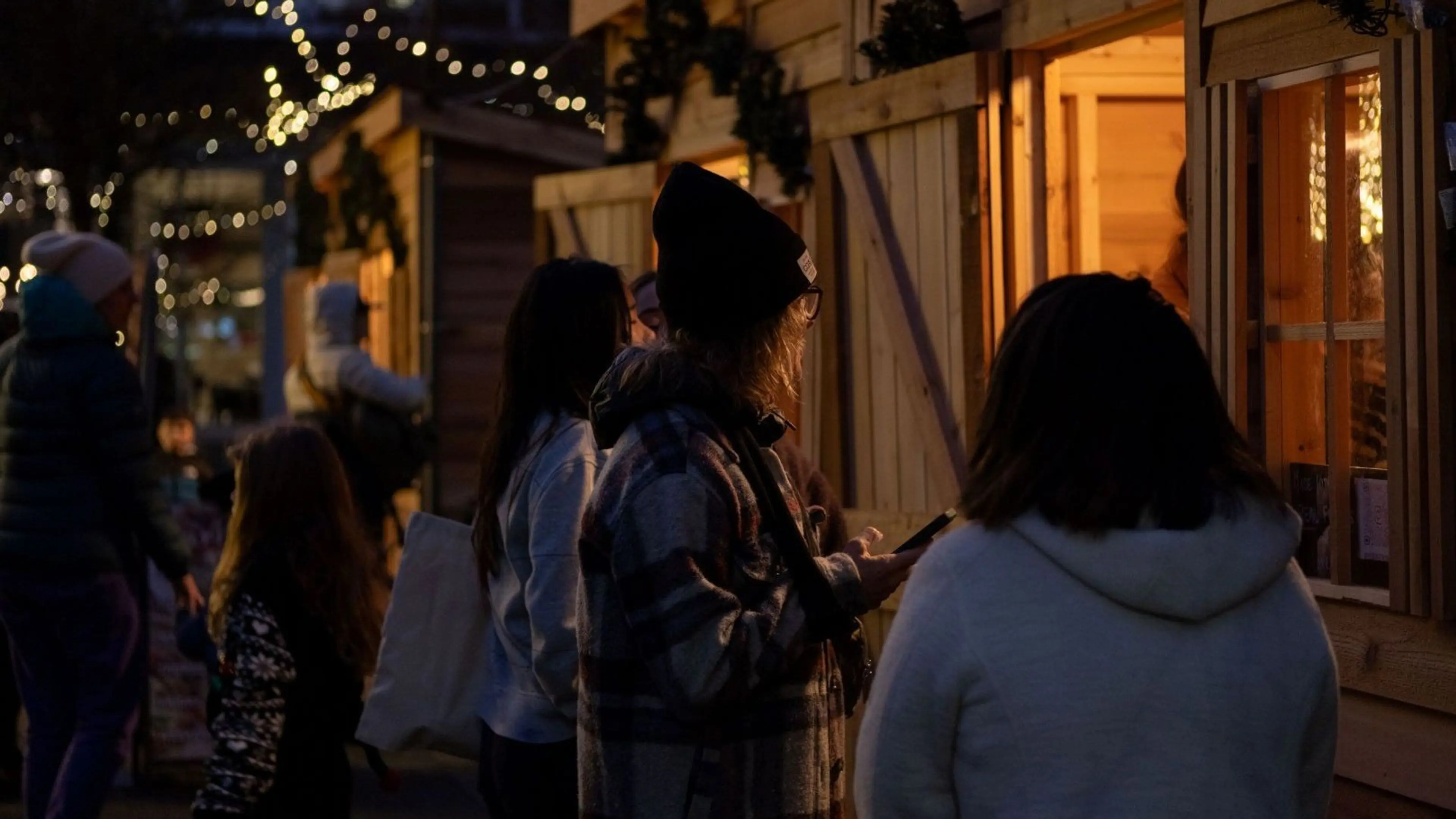 People standing in line at wooden market stalls decorated with lights during evening.