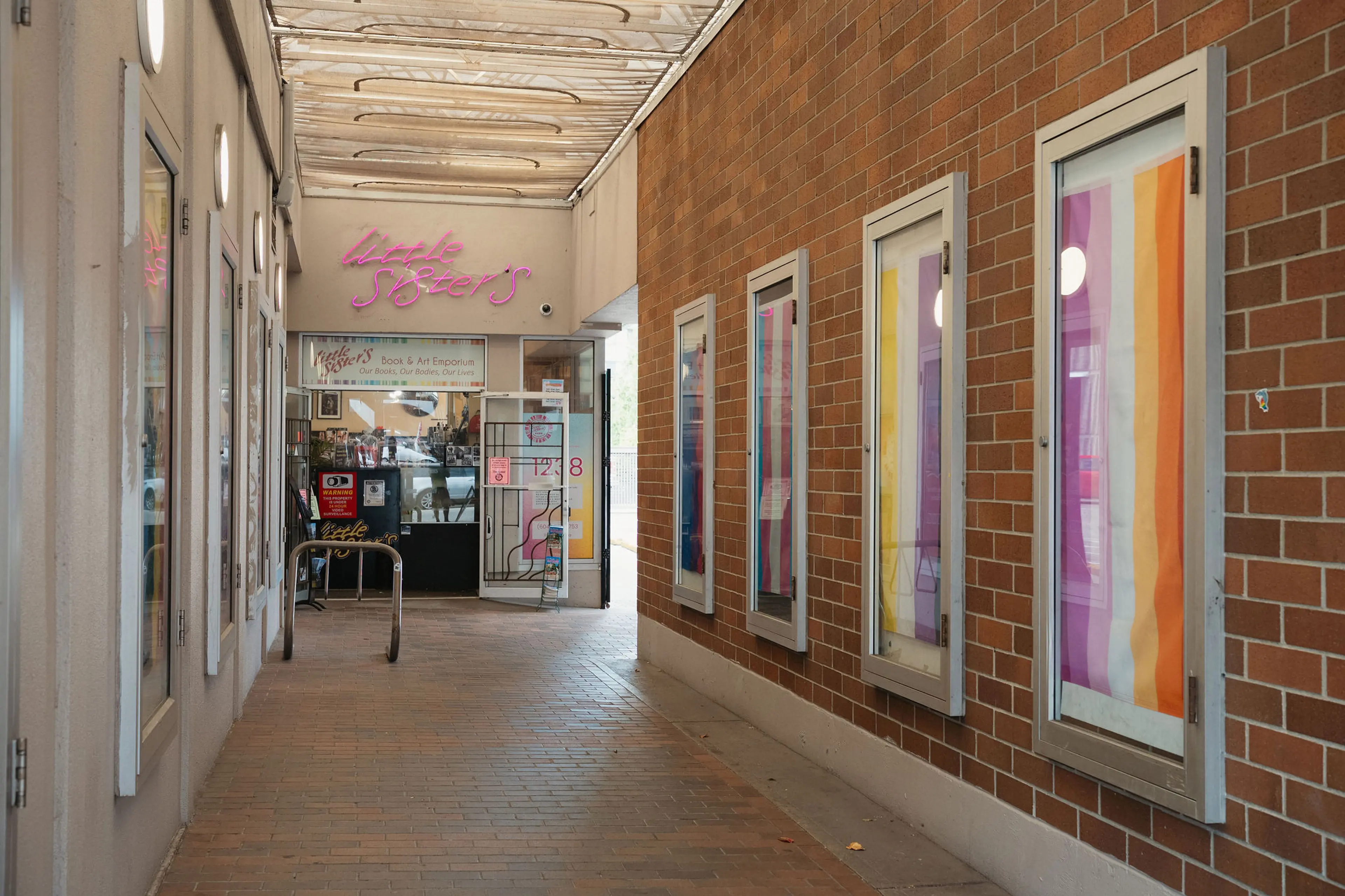 Covered walkway with brick walls and colorful posters on the right, leading to a store with a pink neon sign reading "little sister's".