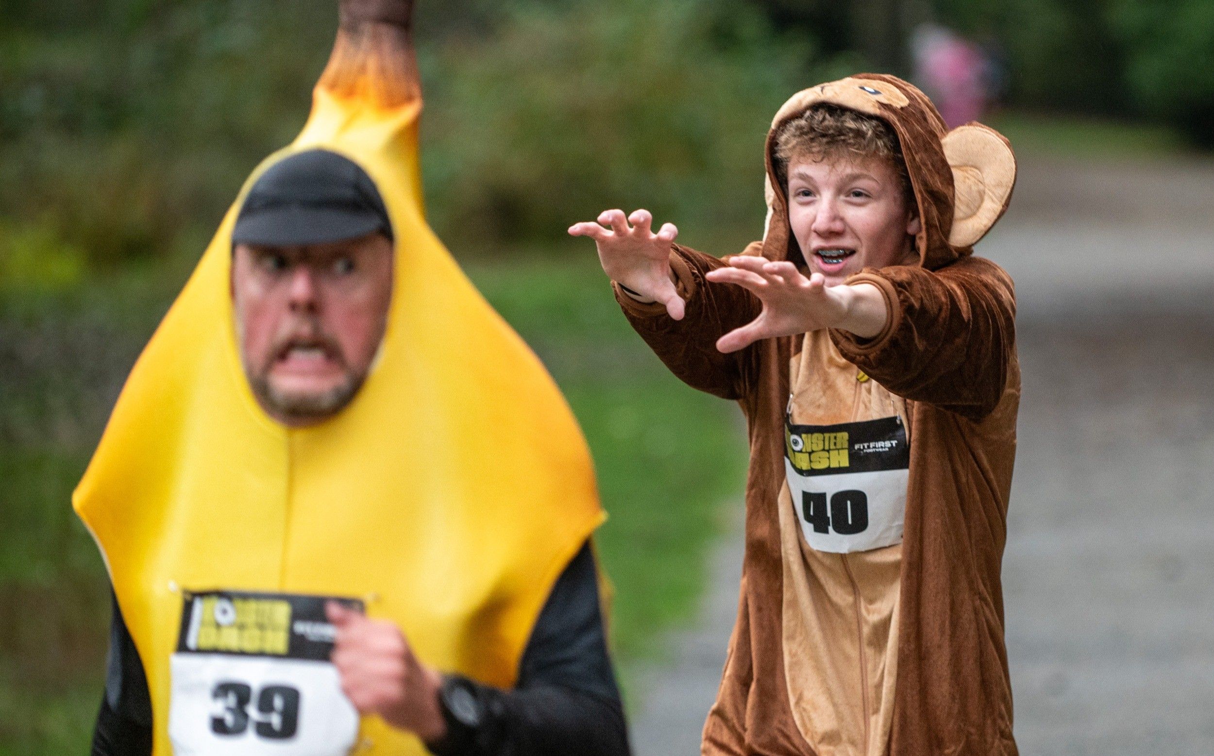 Two people in costumes running outdoors, one in a banana costume and the other in a monkey costume reaching forward.