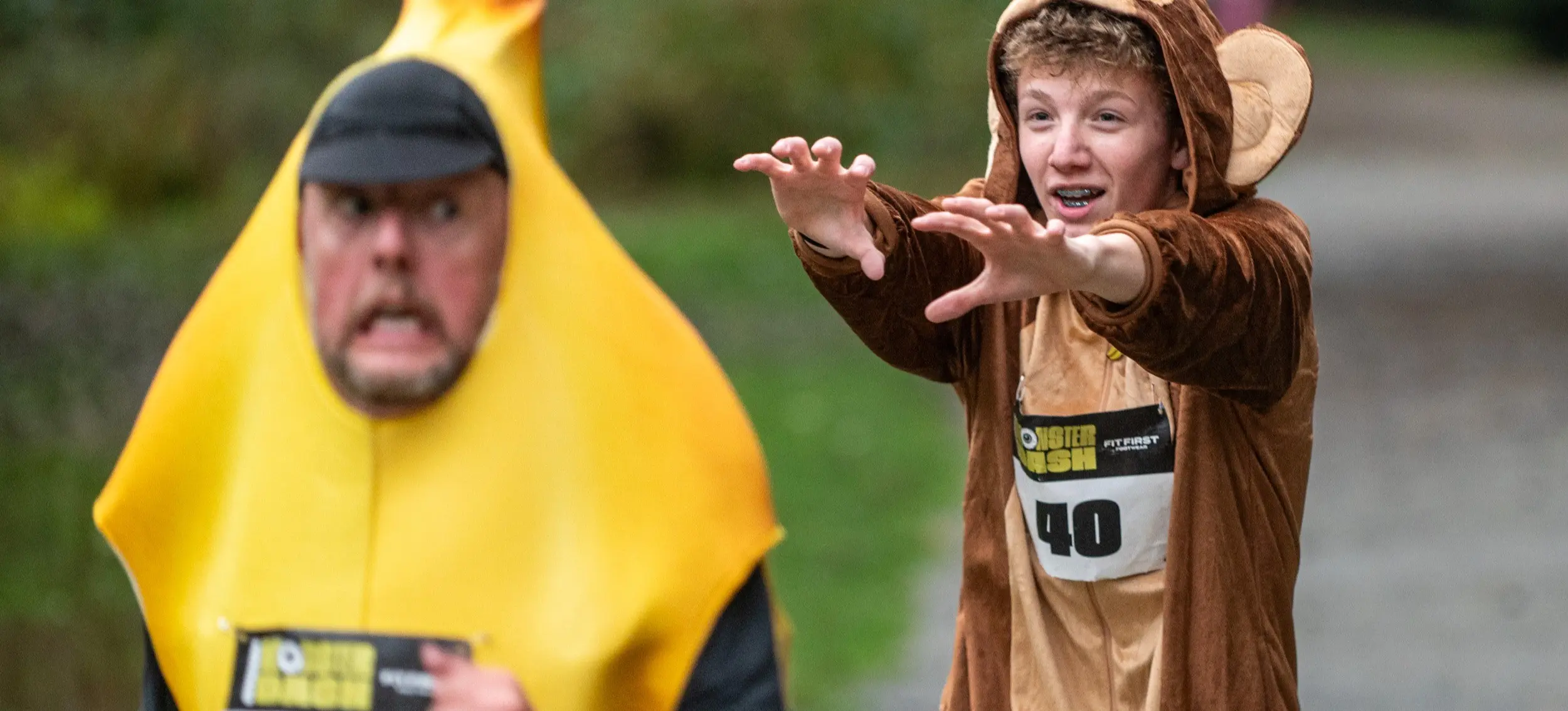 Two people in costumes running outdoors, one in a banana costume and the other in a monkey costume reaching forward.
