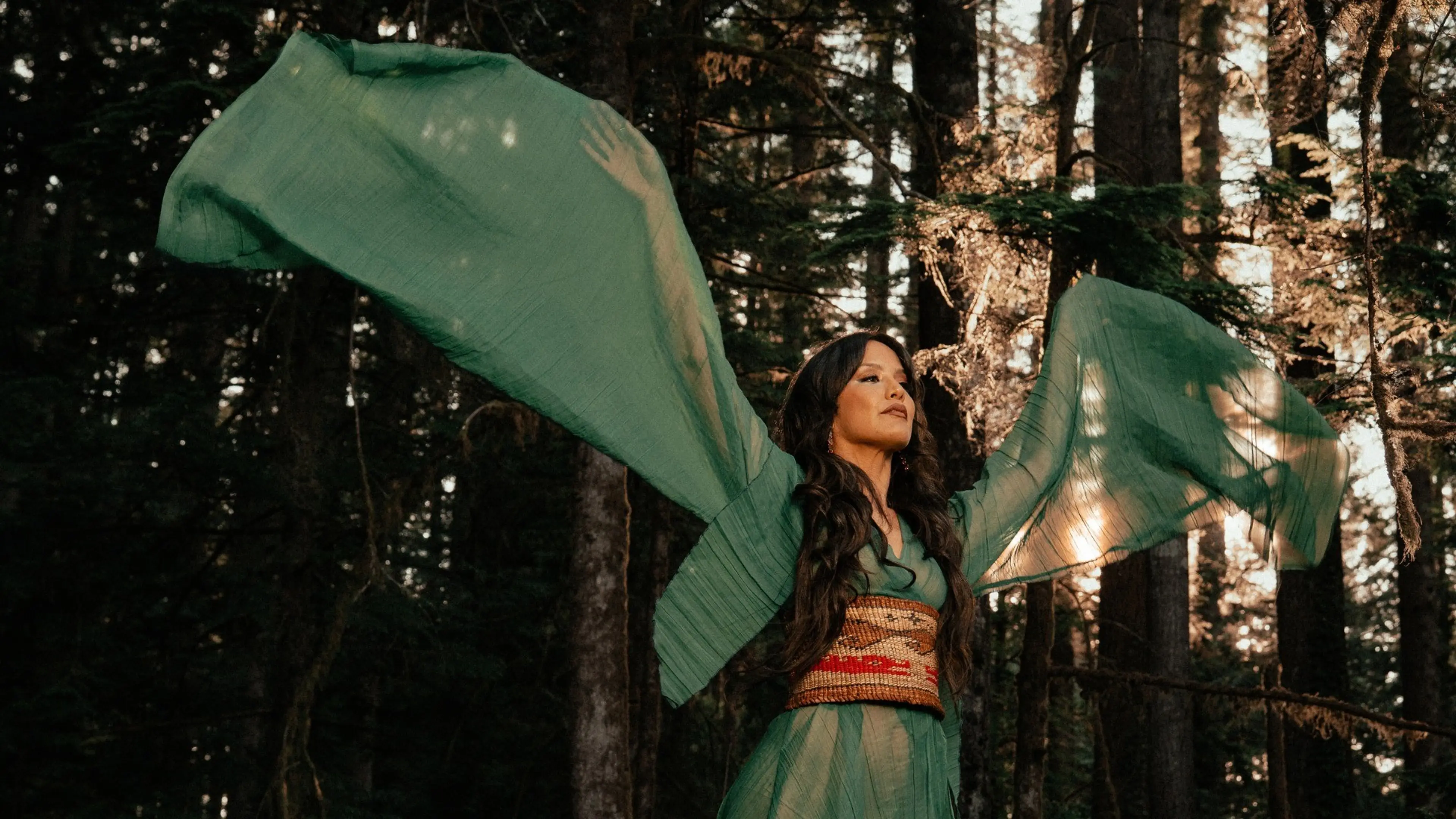 Person in flowing green dress with wide sleeves raised in a forest with sunlight filtering through trees.