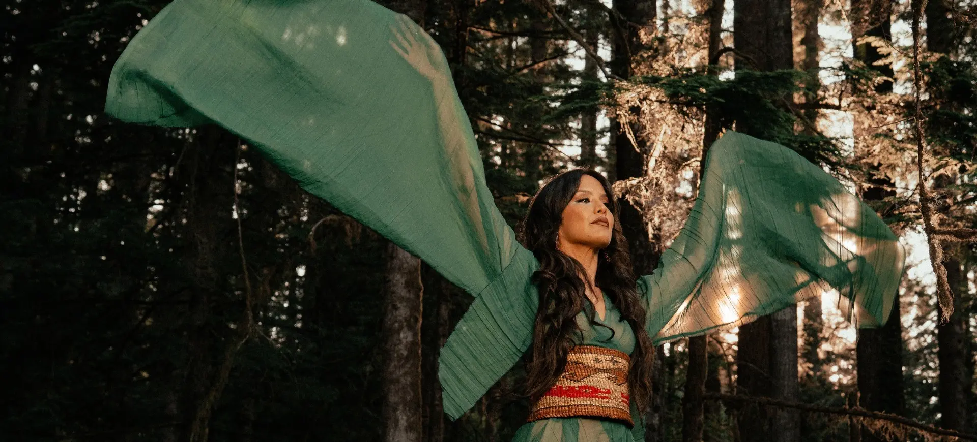 Person in flowing green dress with wide sleeves raised in a forest with sunlight filtering through trees.