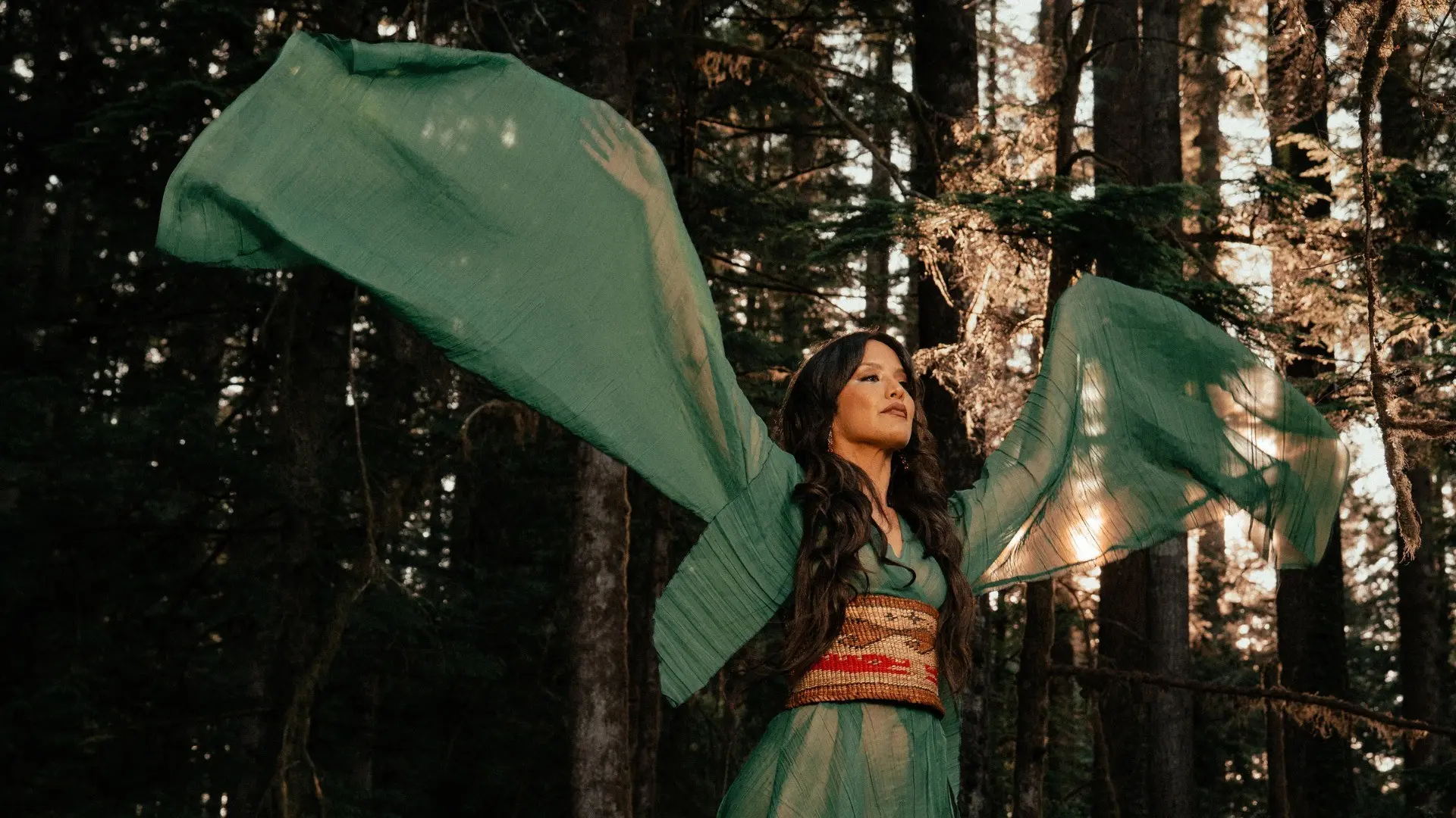 Person in flowing green dress with wide sleeves raised in a forest with sunlight filtering through trees.