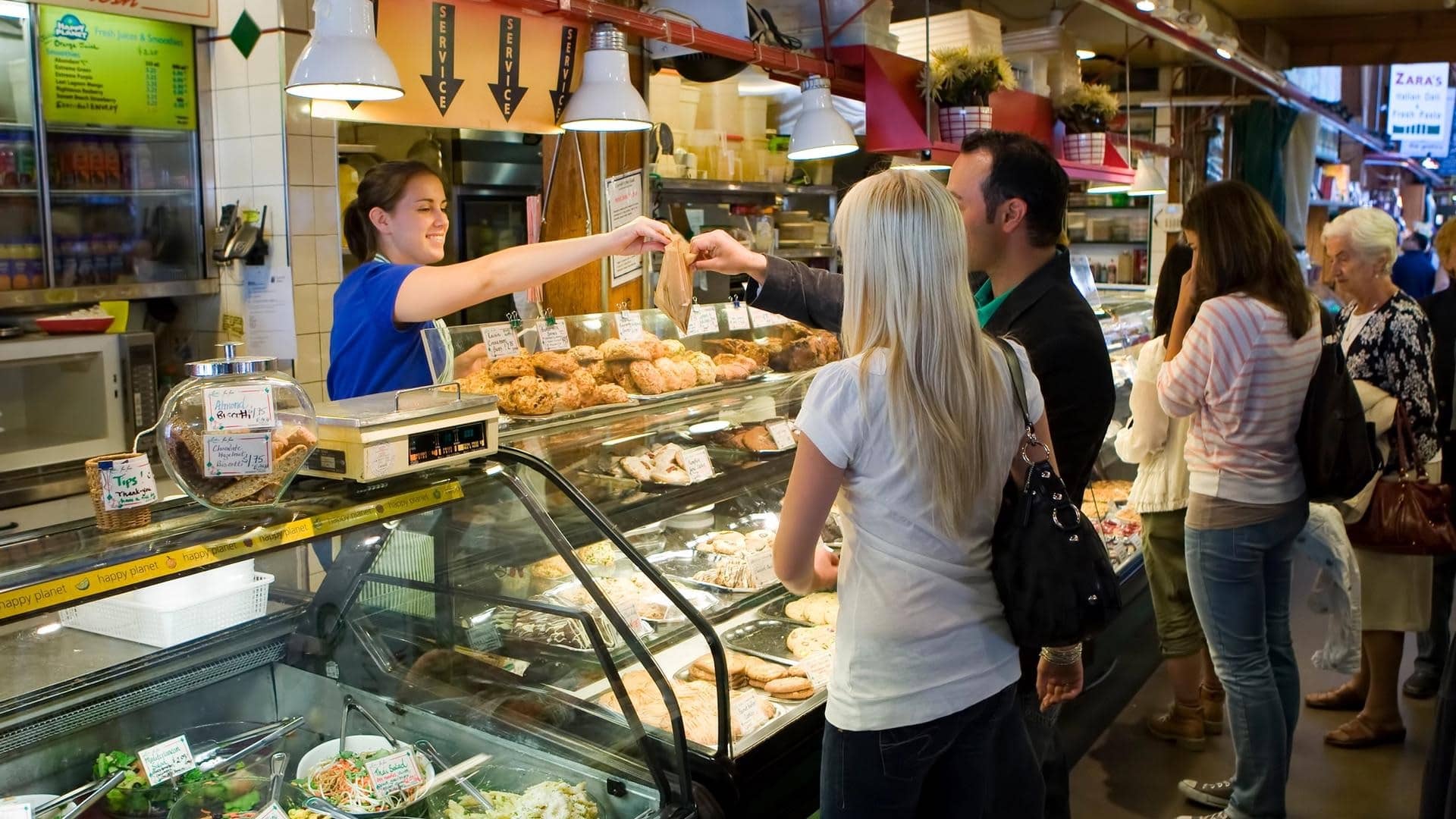 A woman is serving customers at a busy deli and bakery in a public market