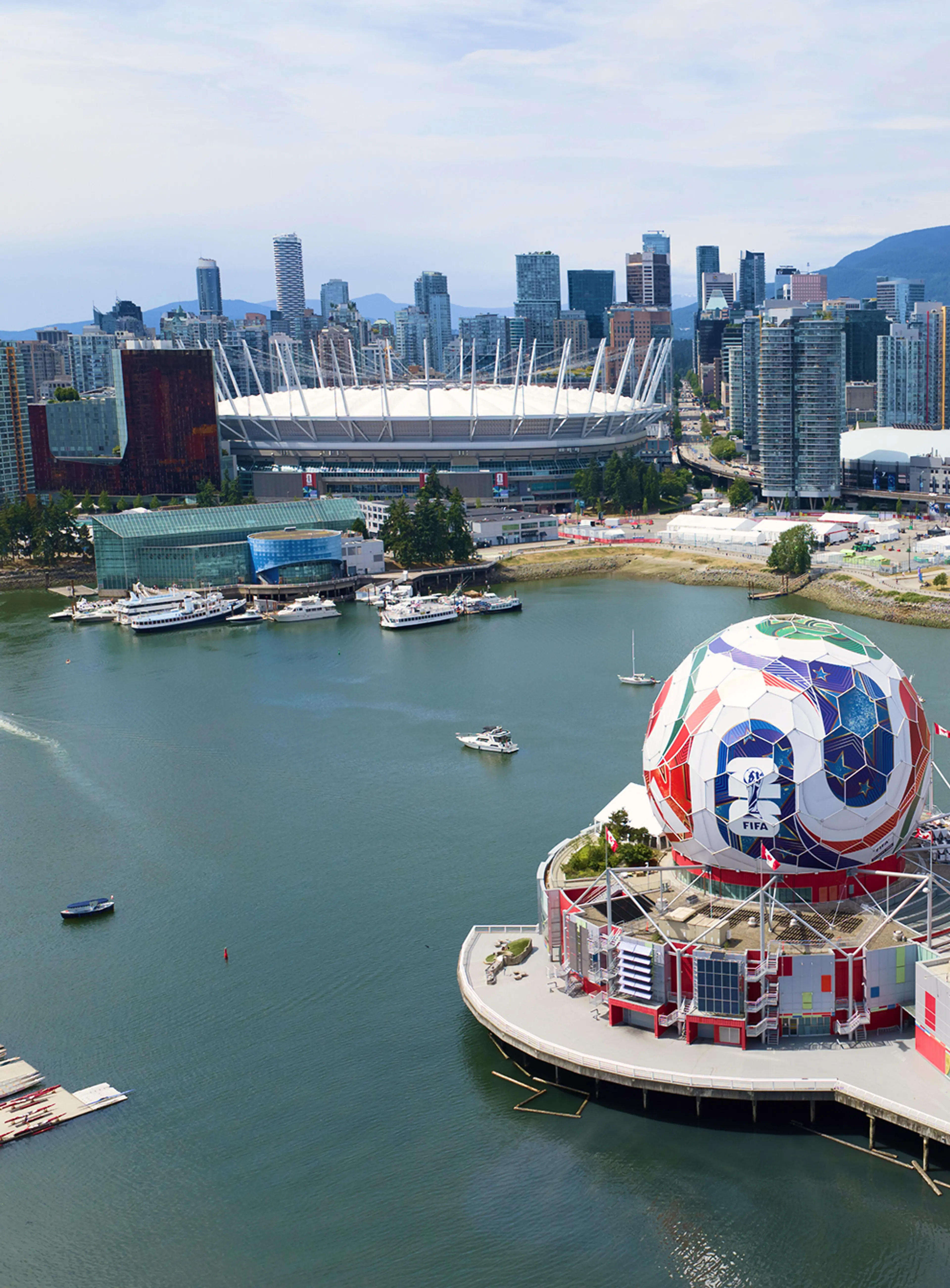 Aerial view of a city skyline with a large stadium, waterfront, and a geodesic dome building under a partly cloudy sky.