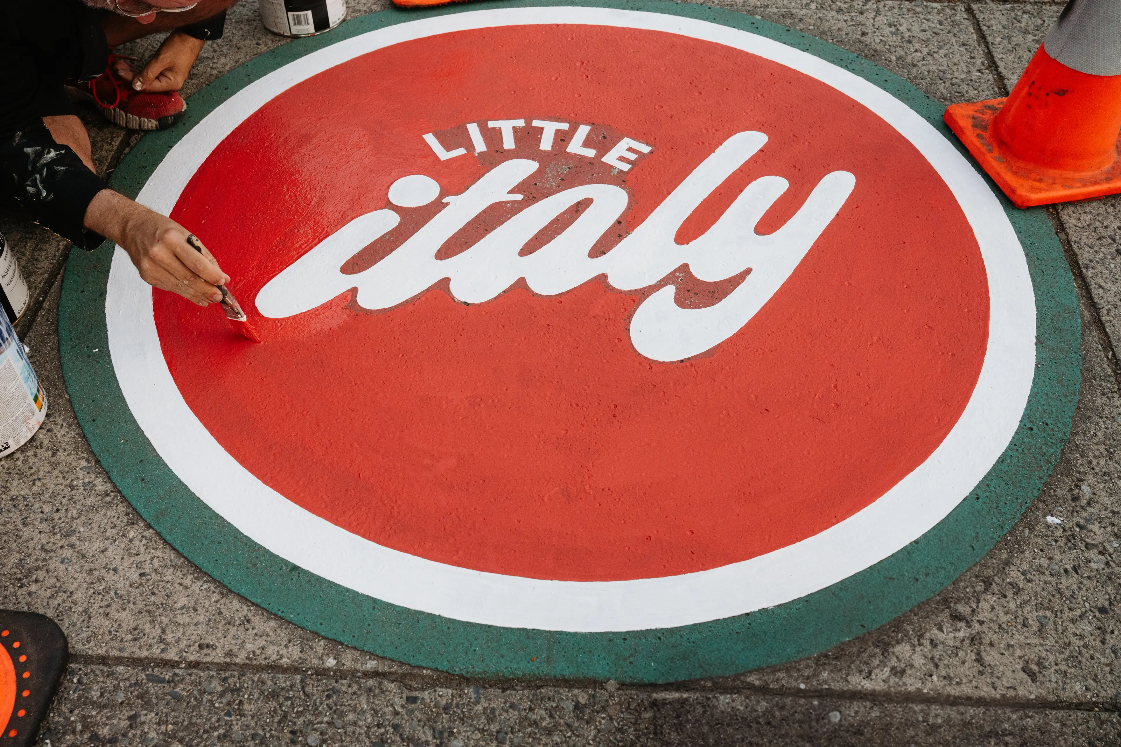 Person painting a circular street sign with red, white, and green colors that reads 'Little Italy'.