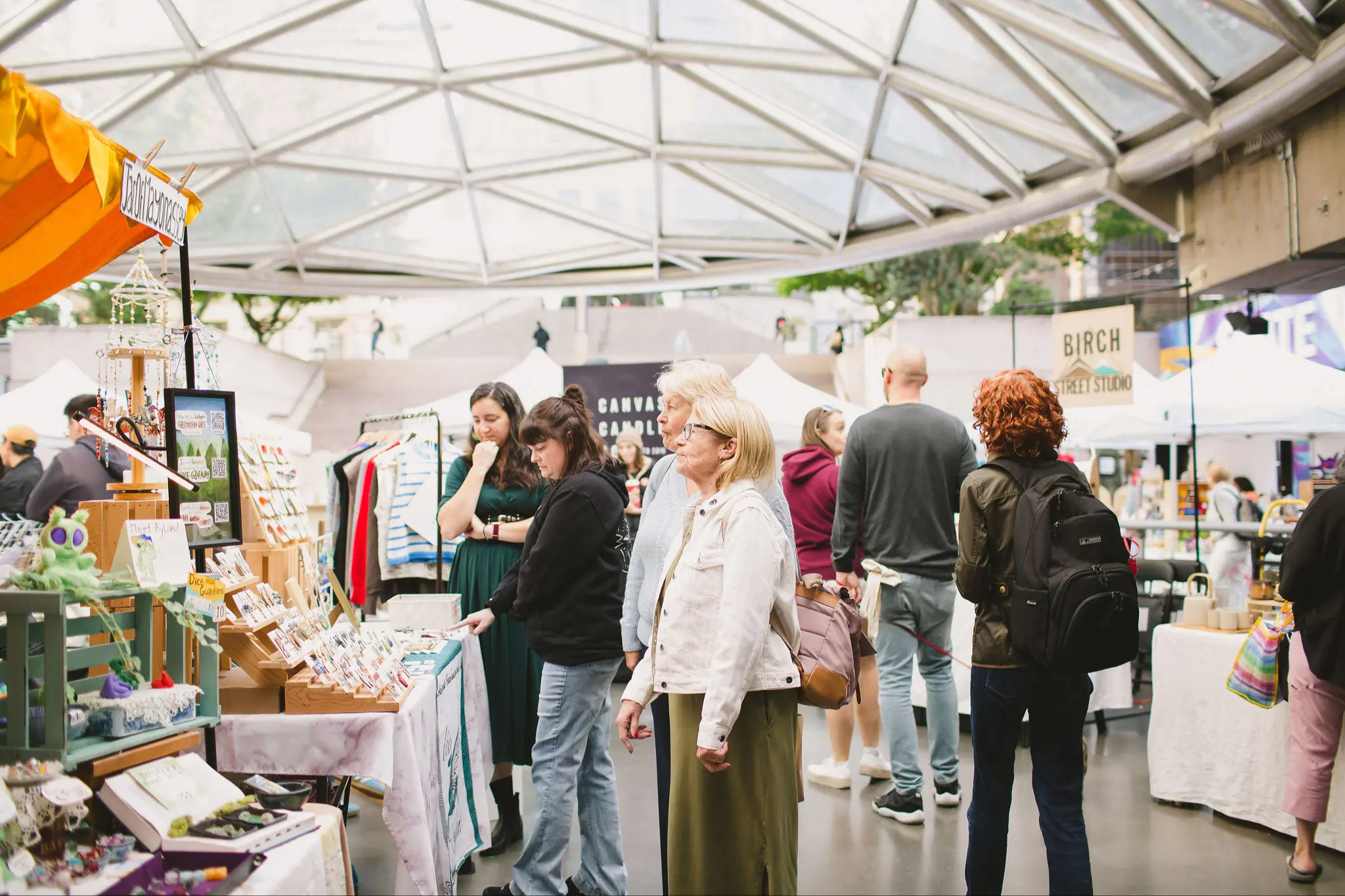 People browsing various stalls with crafts and goods under a glass canopy at an indoor market.