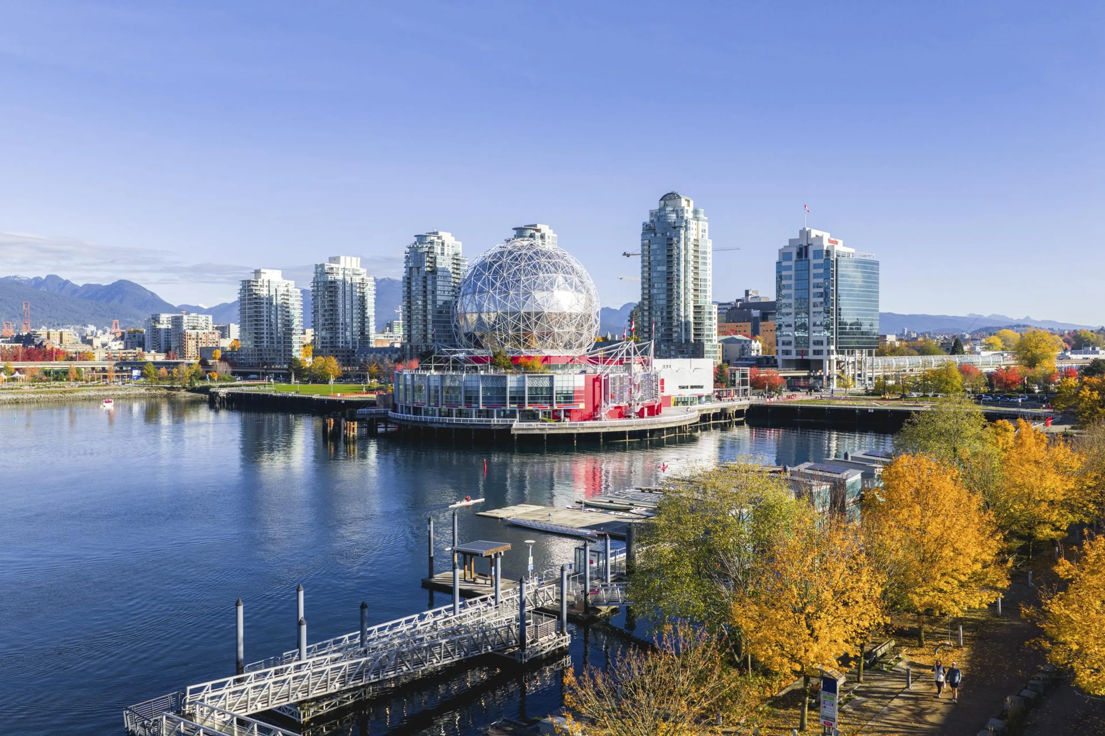 Waterfront cityscape with a geodesic dome building, high-rise buildings, and autumn trees along a walkway.