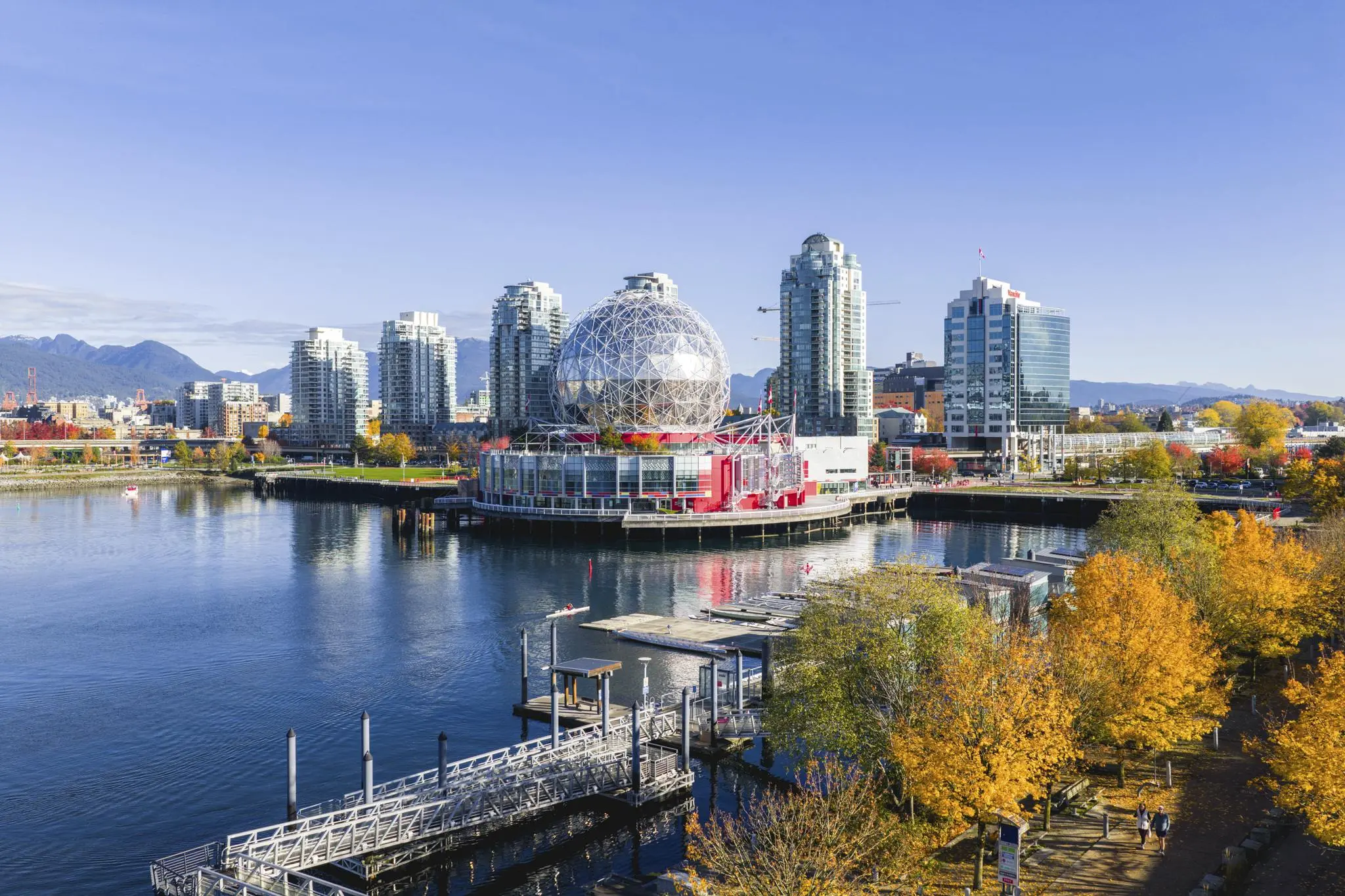 Waterfront cityscape with a geodesic dome building, high-rise buildings, and autumn trees along a walkway.