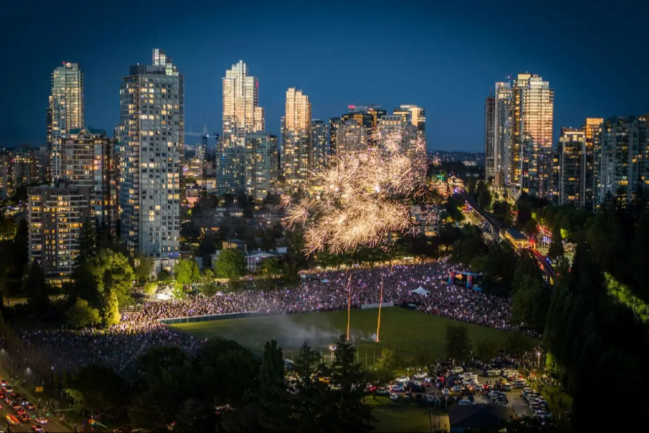 Fireworks display over a crowded park surrounded by tall city buildings at night.