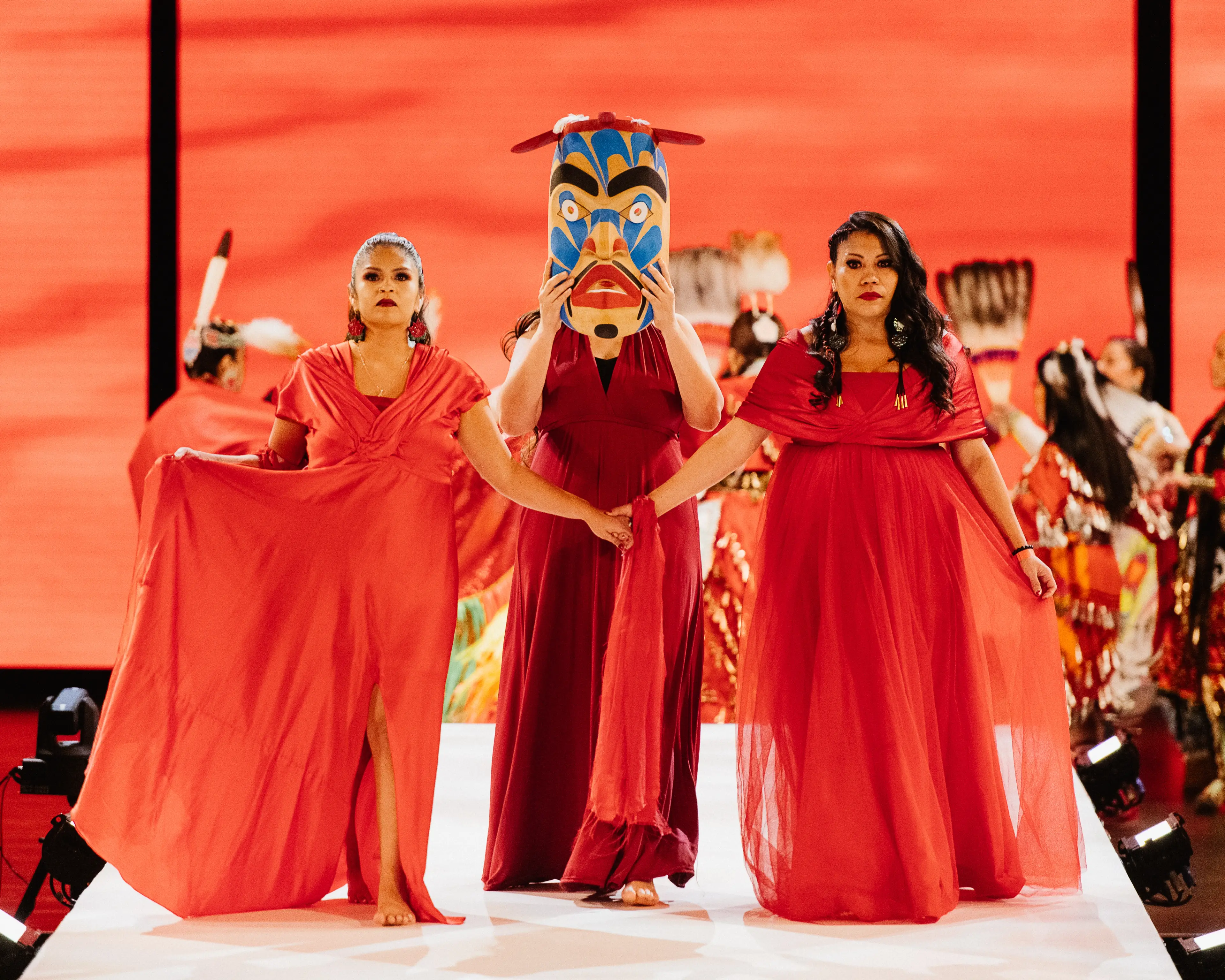 Three individuals, dressed in red, on the runway for Red Dress Night (one holds a mask in front of their face)