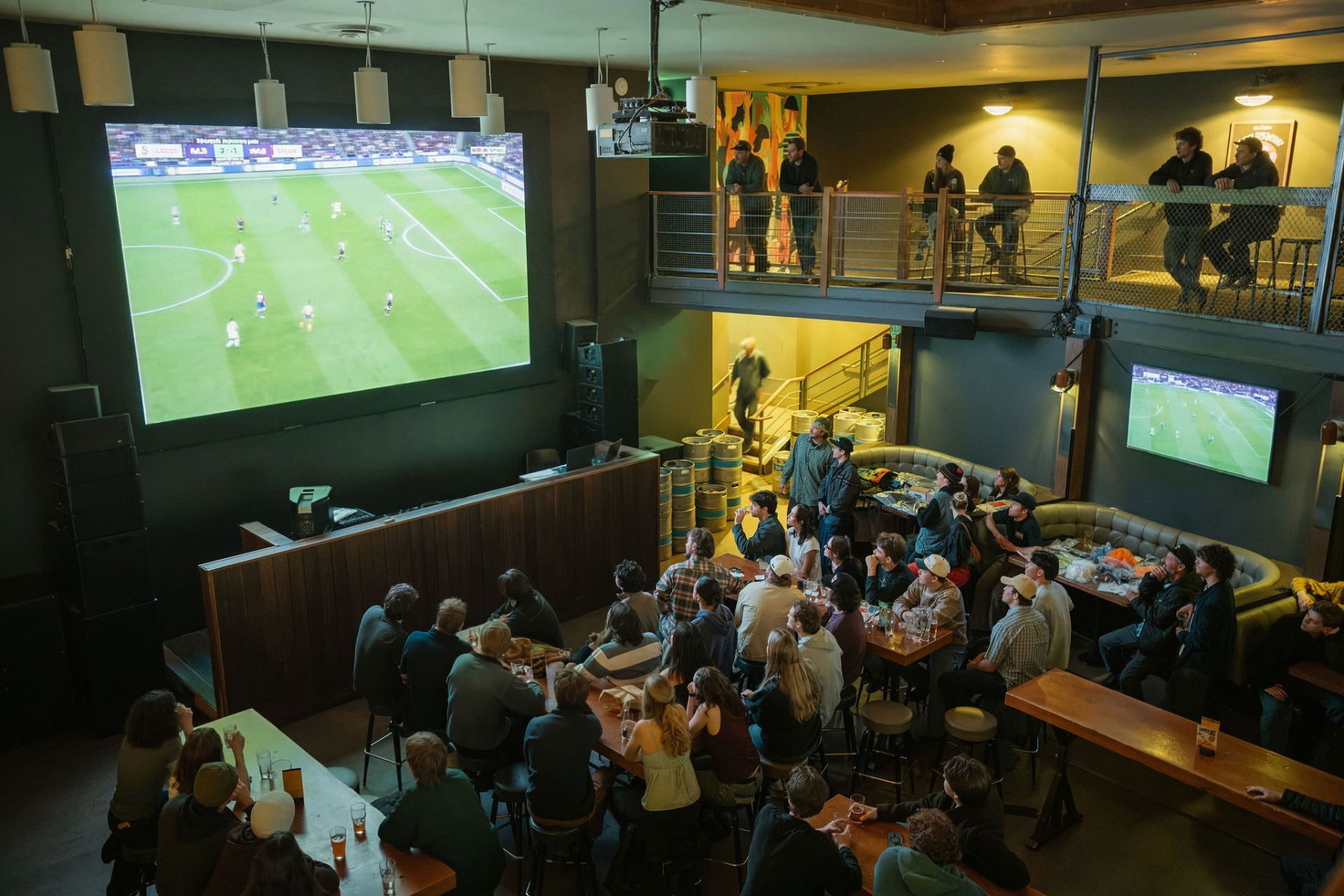 Crowd of people watching a soccer match on a large screen in a dimly lit bar with a balcony and smaller screen.