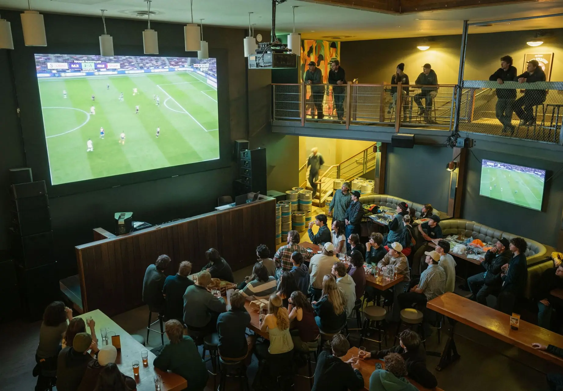 Crowd of people watching a soccer match on a large screen in a dimly lit bar with a balcony and smaller screen.