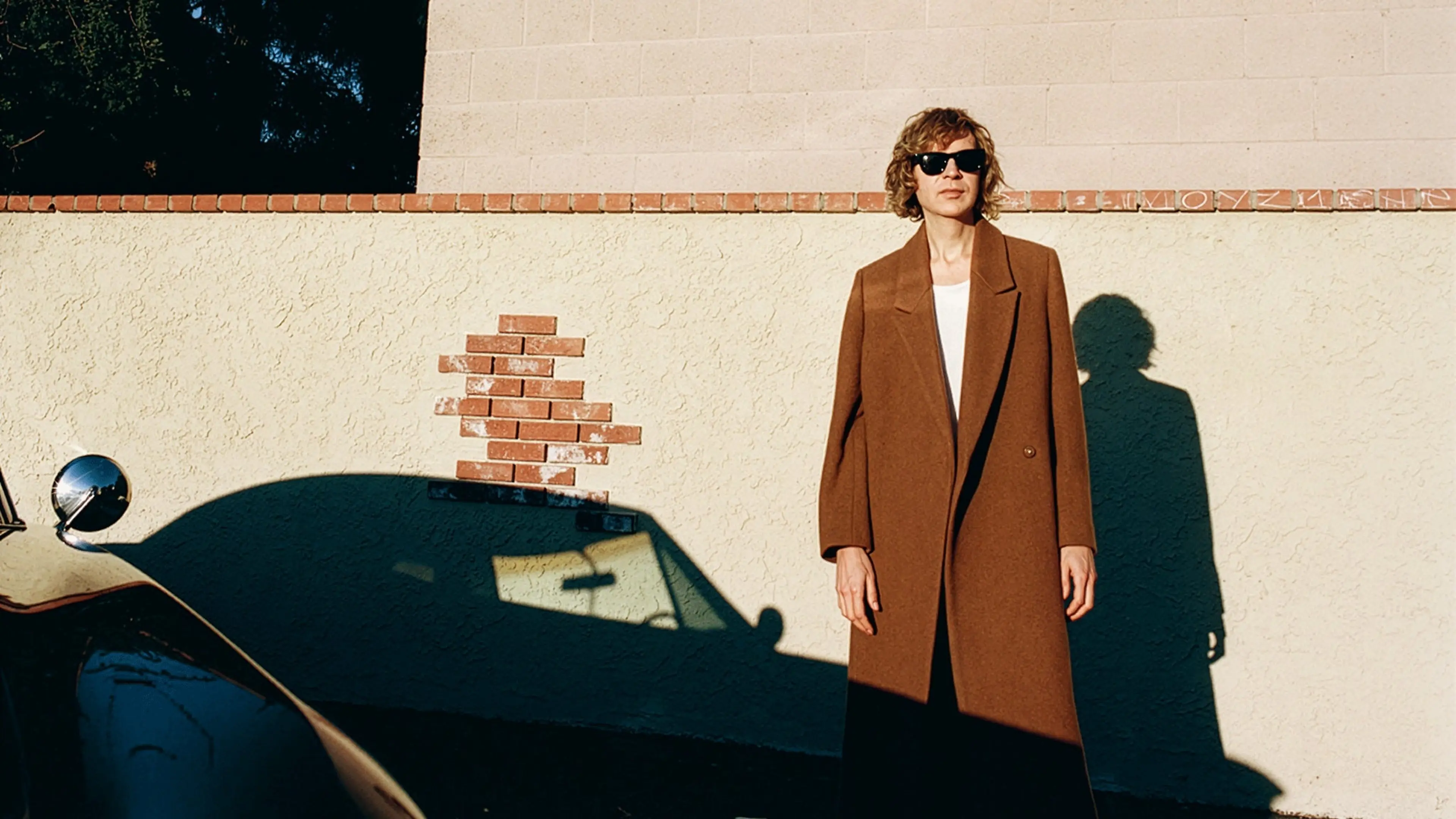 Person wearing brown coat and sunglasses standing against a beige wall with shadow of a car and the person visible.
