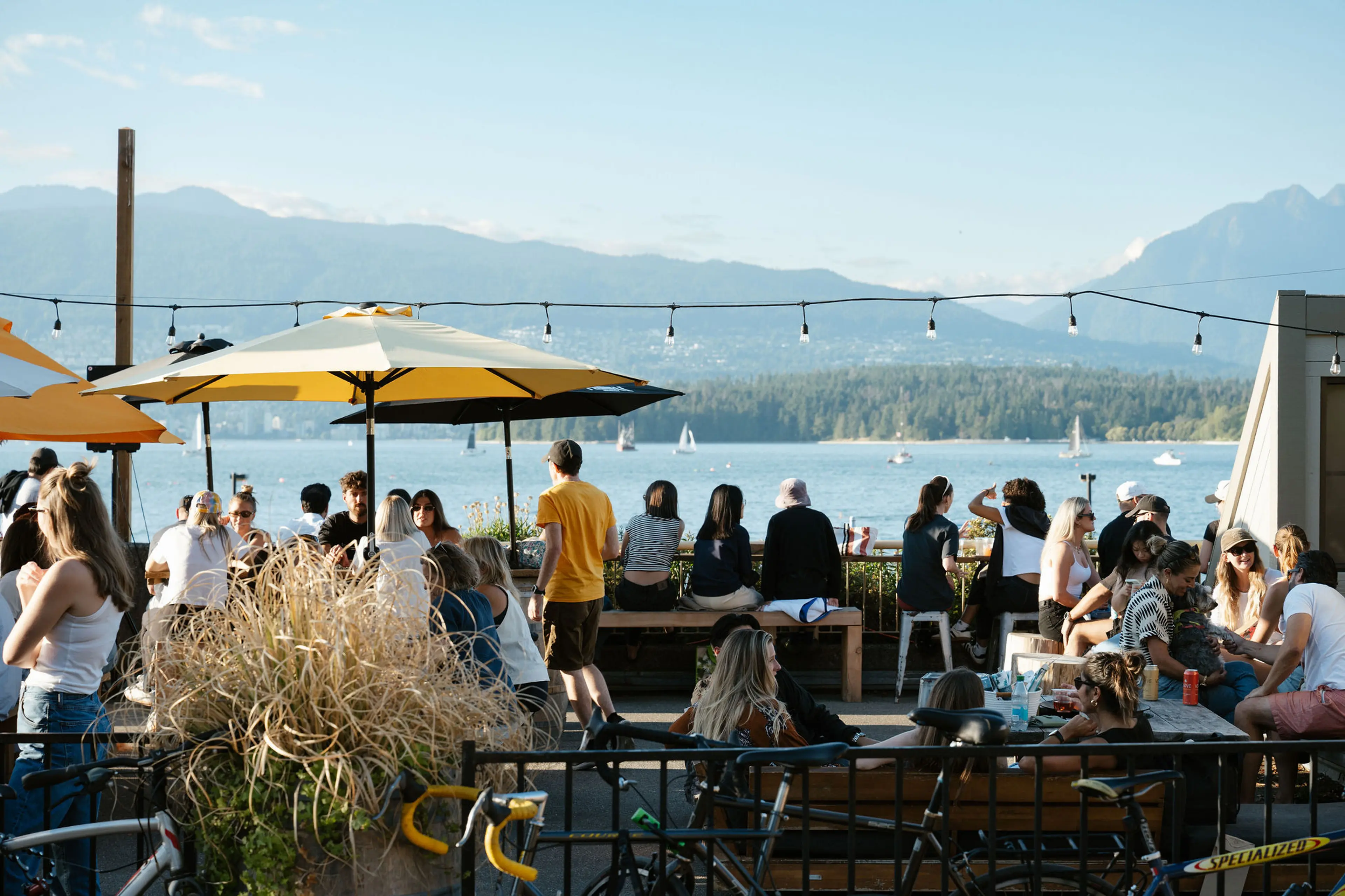 People sitting and standing at a patio by a waterfront with mountains and sailboats in the background.