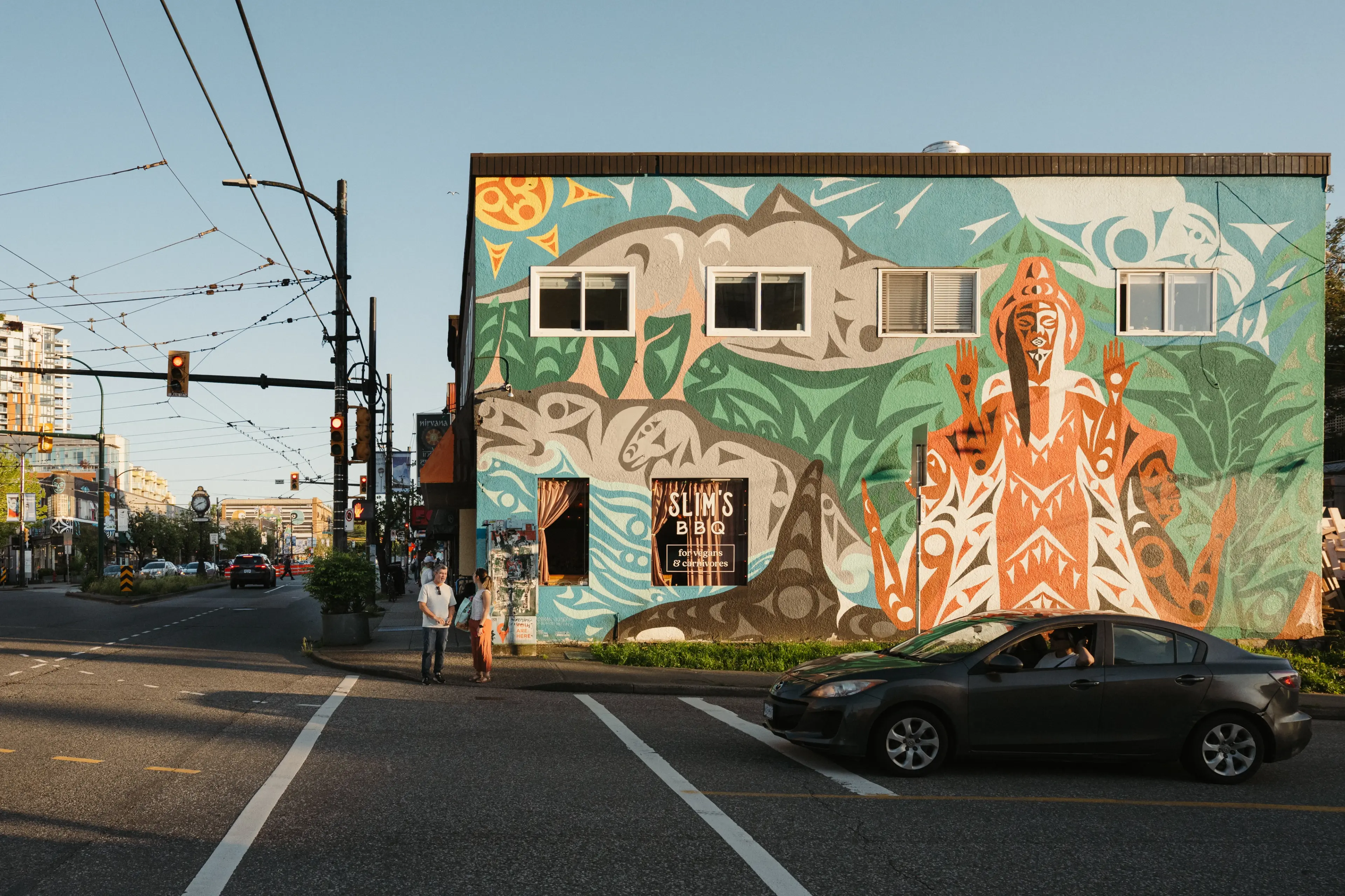 Building with colorful mural featuring abstract animal and human figures, with a sign for Slim's BBQ, at a street intersection with traffic lights and pedestrians.