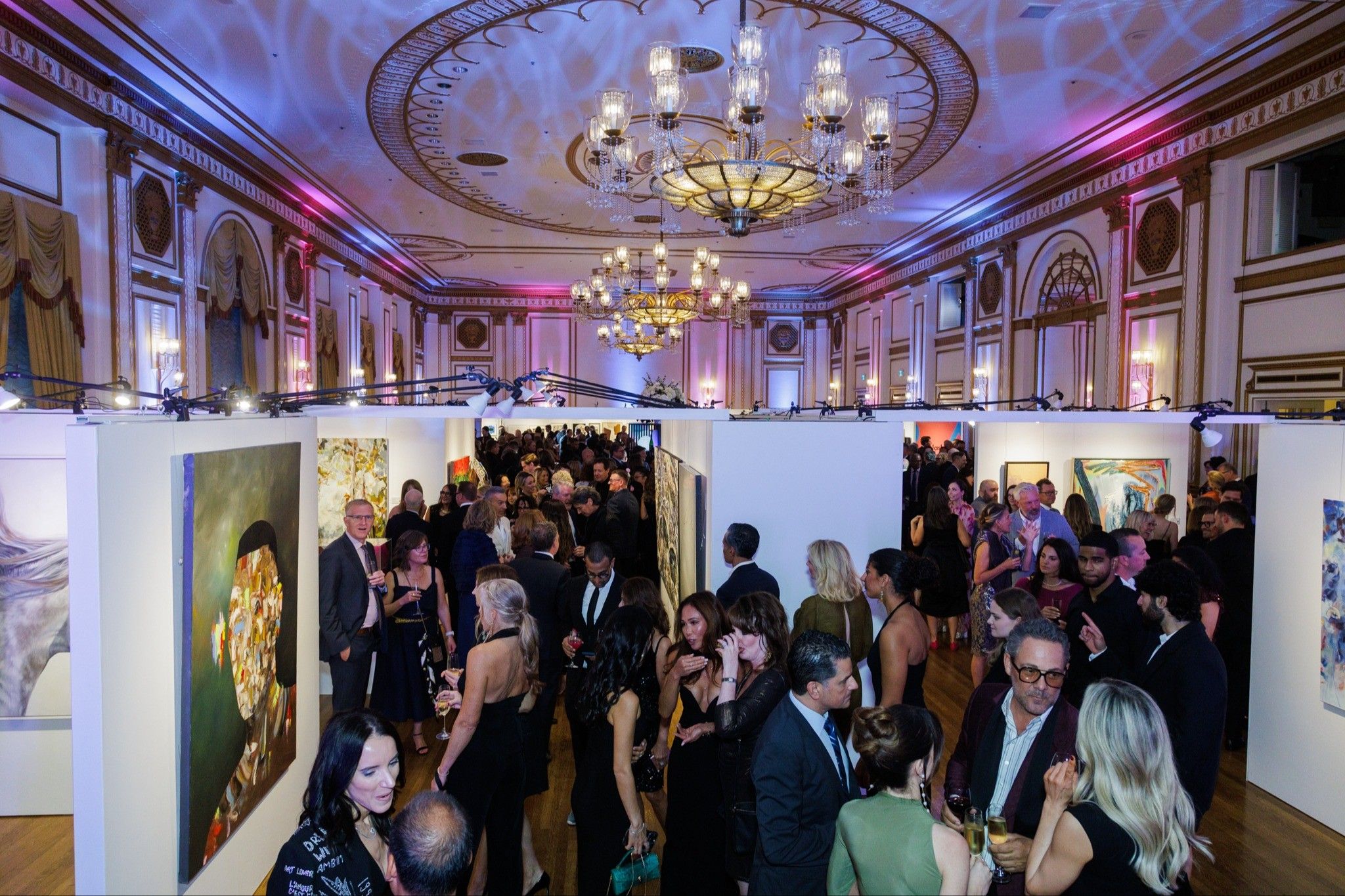 Crowded art gallery event with people dressed formally, viewing paintings on white walls under ornate chandeliers.