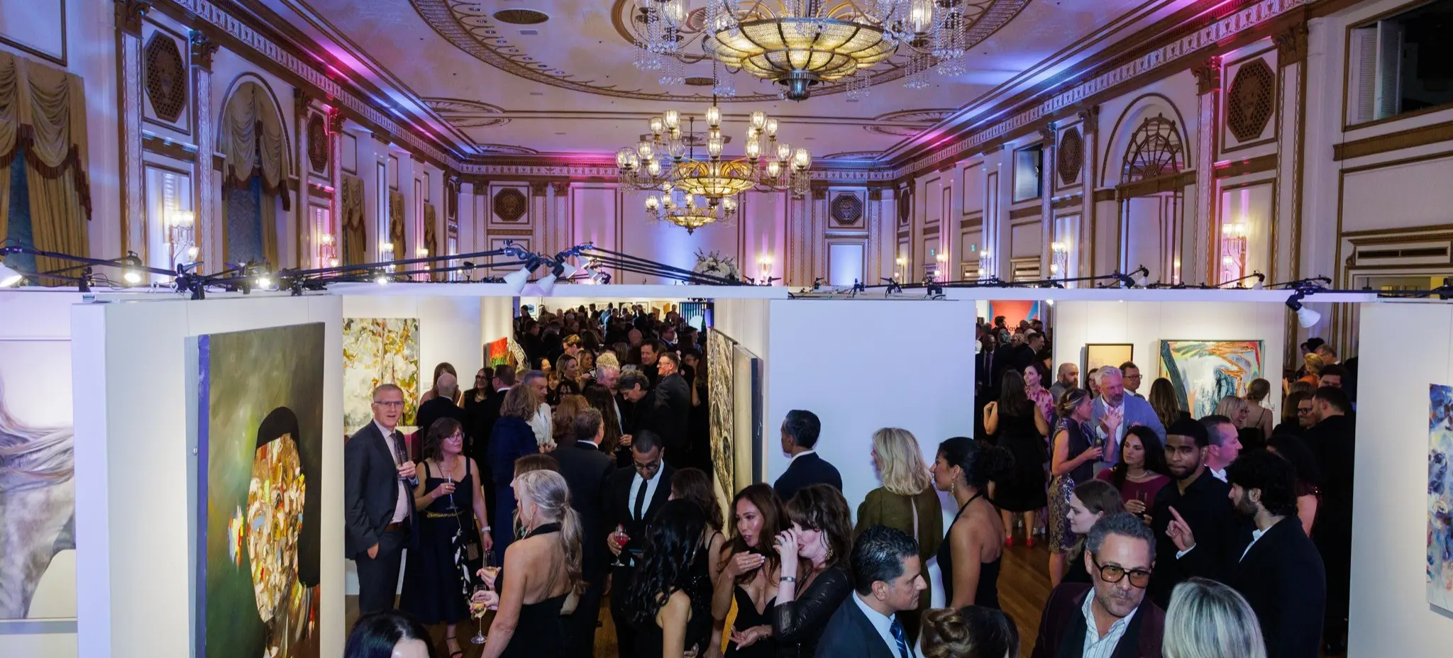 Crowded art gallery event with people dressed formally, viewing paintings on white walls under ornate chandeliers.