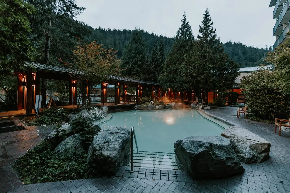 Outdoor hot spring pool surrounded by large rocks, trees, and a wooden walkway with warm lights at dusk.