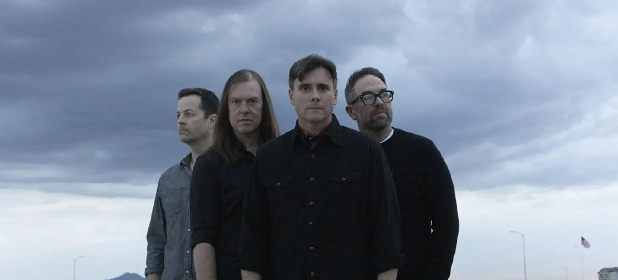 Four people standing outdoors under a cloudy sky, looking in different directions, with a distant American flag visible.