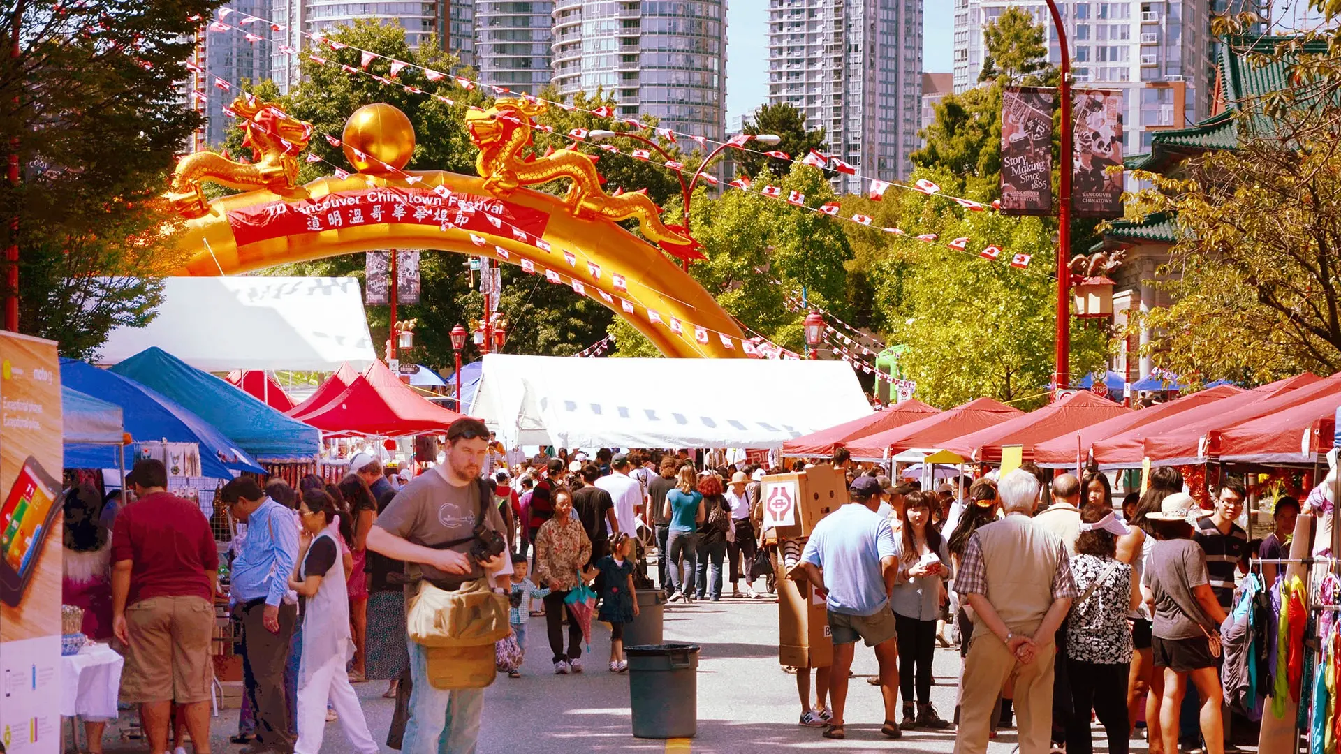 Crowd walking through outdoor market stalls under a large yellow arch with dragons and Canadian flags in a city setting.