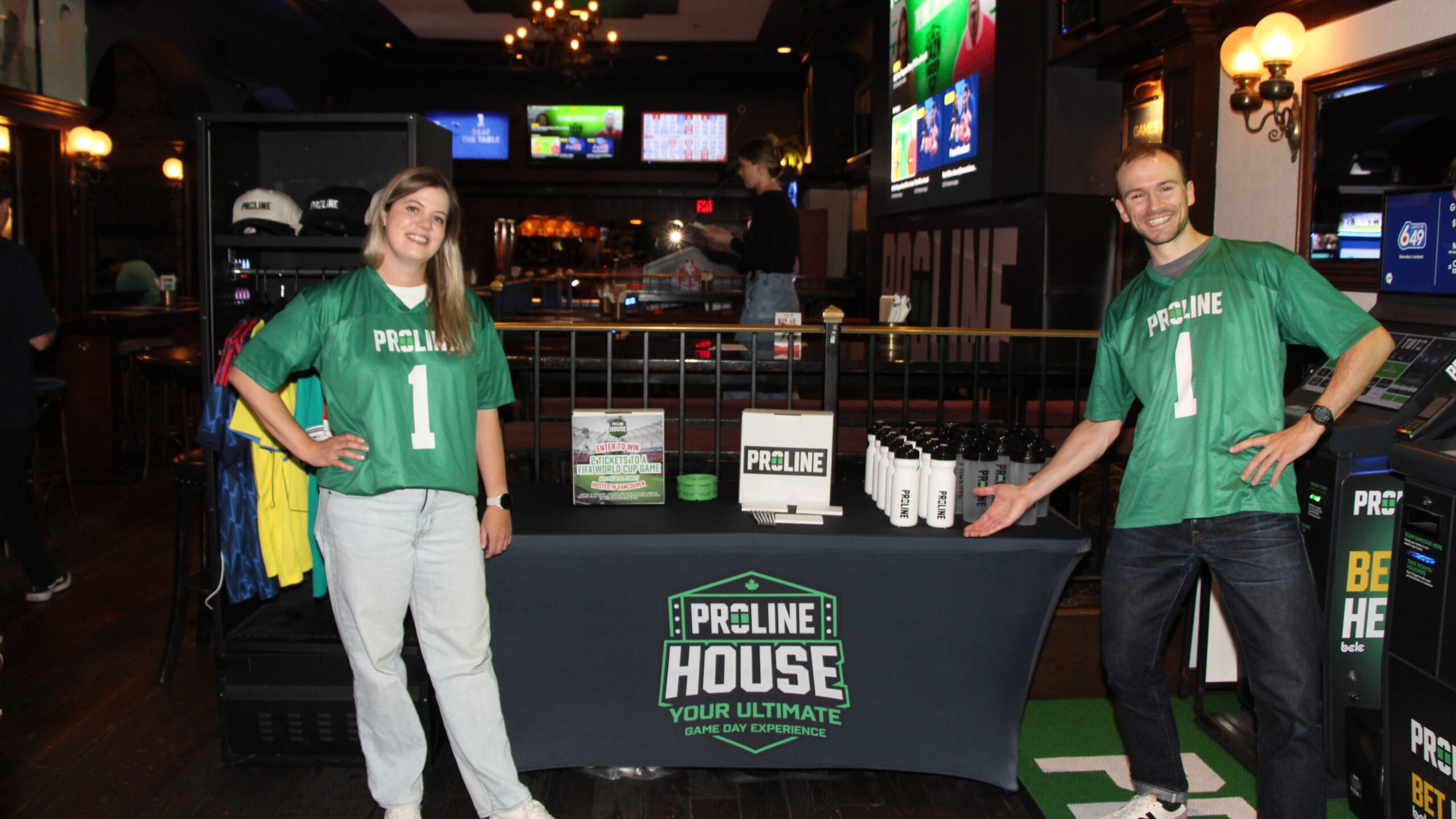 Two people in green Proline jerseys standing beside a table with Proline House promotional items in a bar setting.