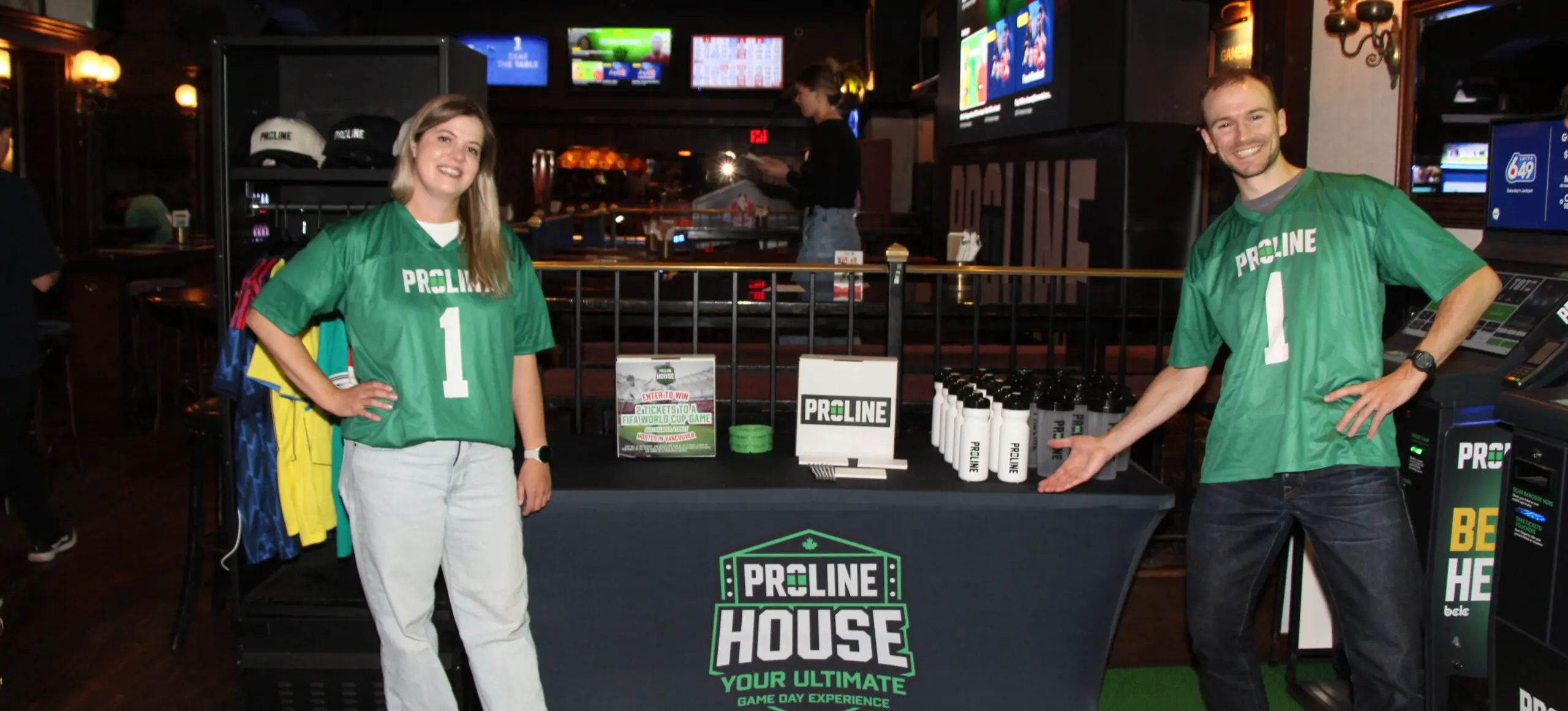Two people in green Proline jerseys standing beside a table with Proline House promotional items in a bar setting.