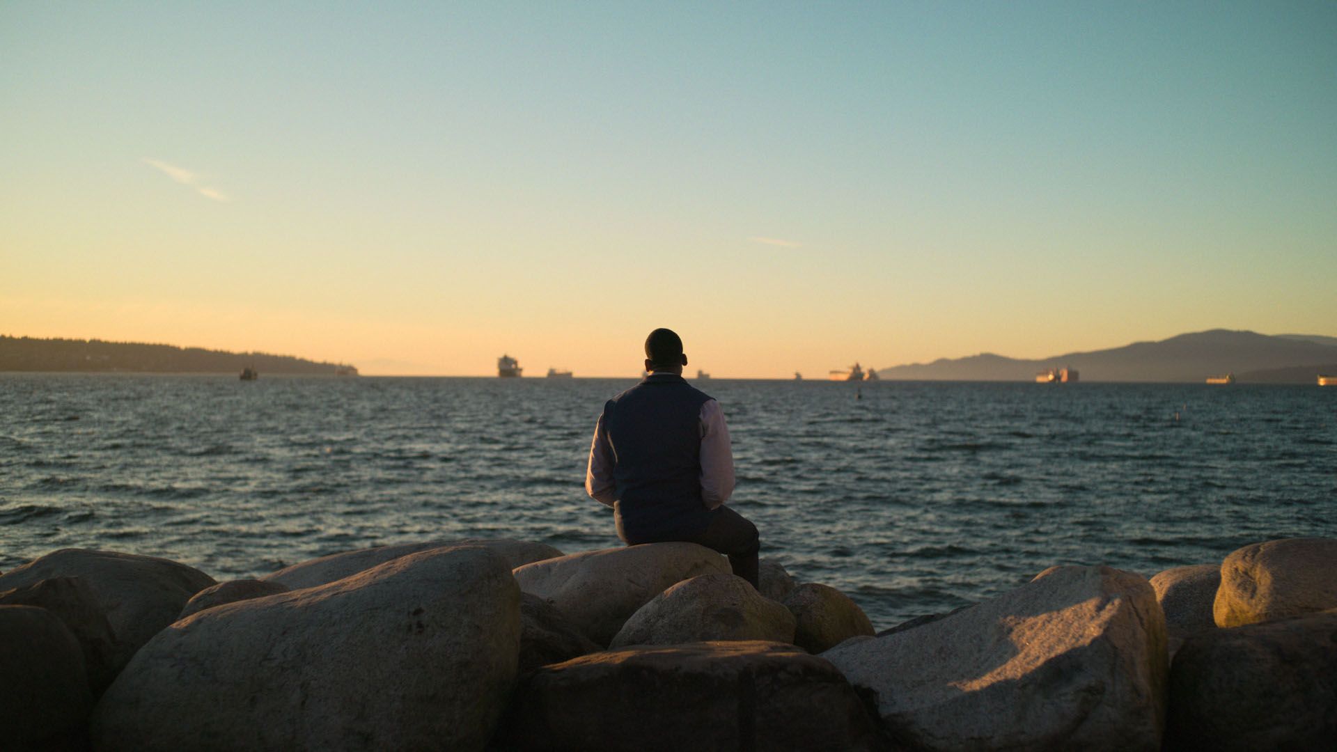 Person sitting on large rocks facing the ocean at sunset with ships on the horizon.