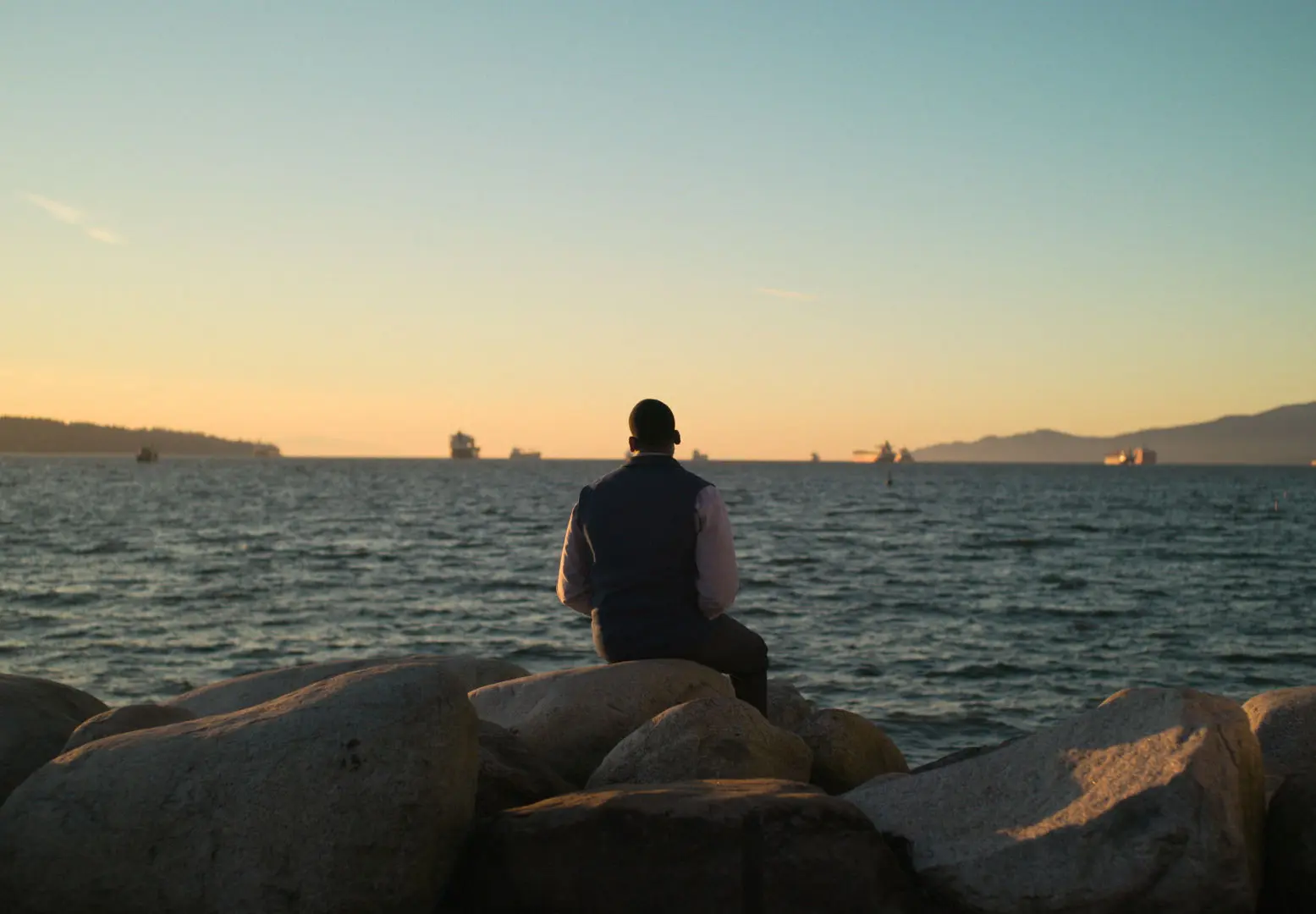 Person sitting on large rocks facing the ocean at sunset with ships on the horizon.