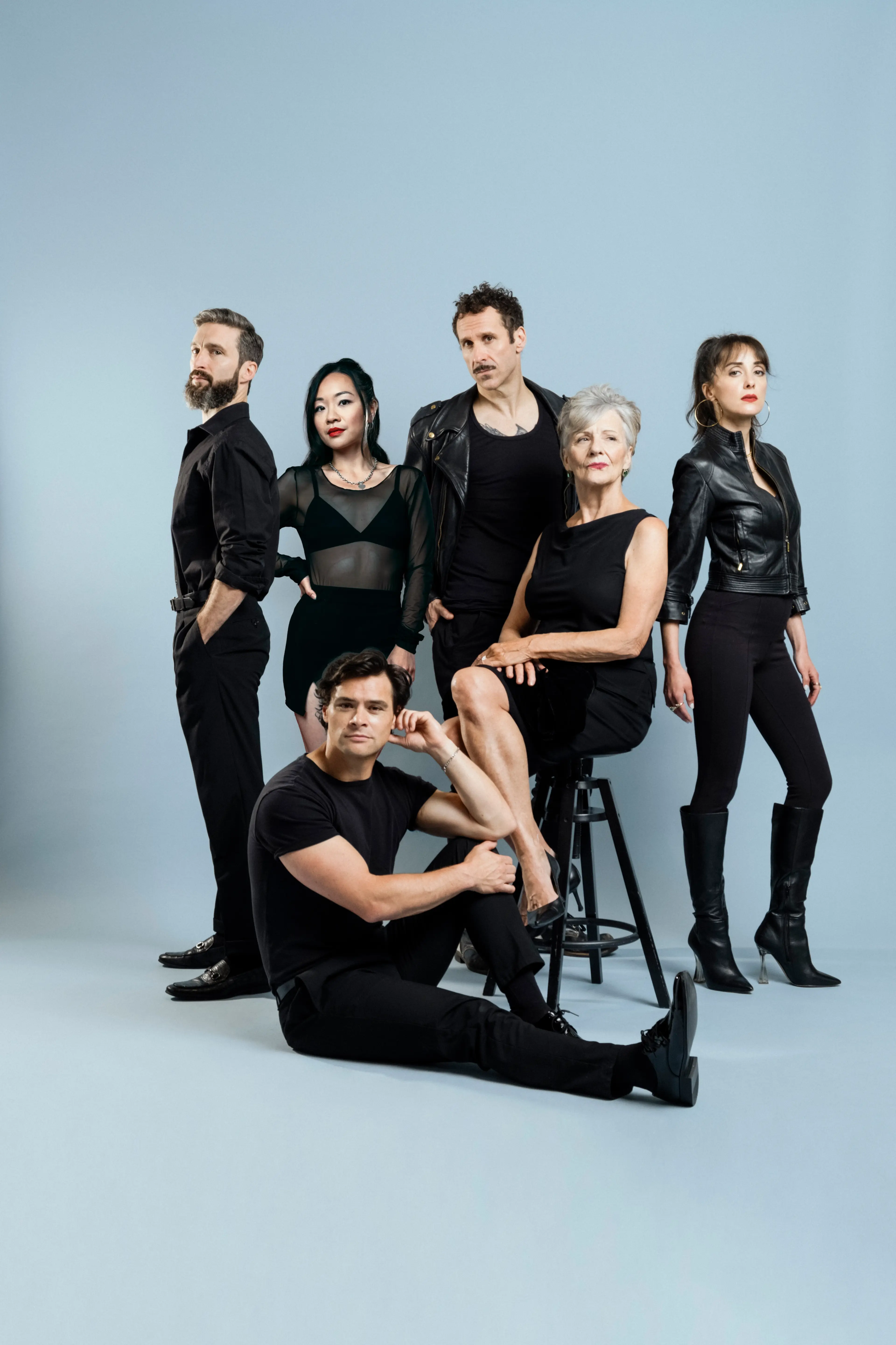 Group of six people dressed in black posing against a light blue background, one seated on a stool, one sitting on the floor, others standing.