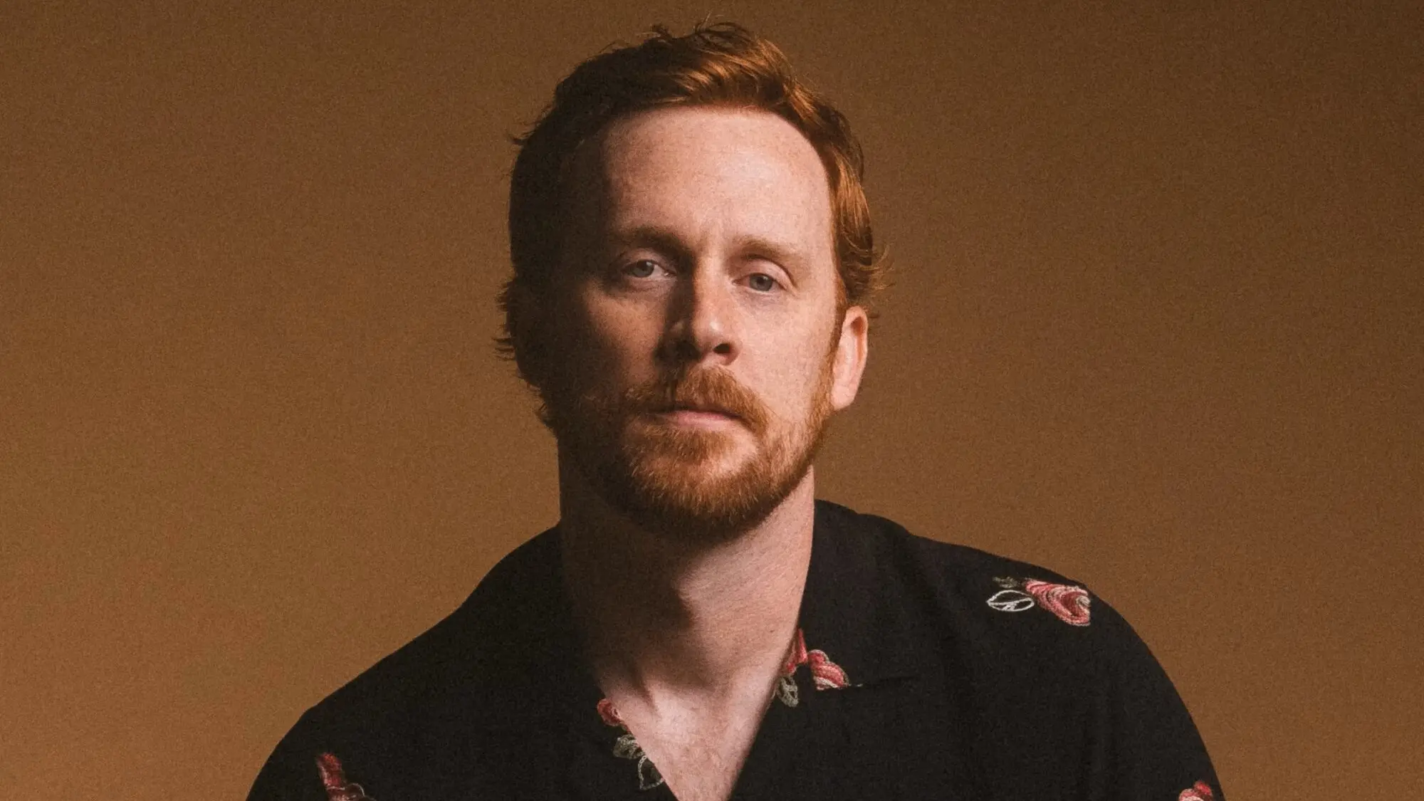 Person with red hair and beard wearing a black shirt with floral patterns against a brown background.