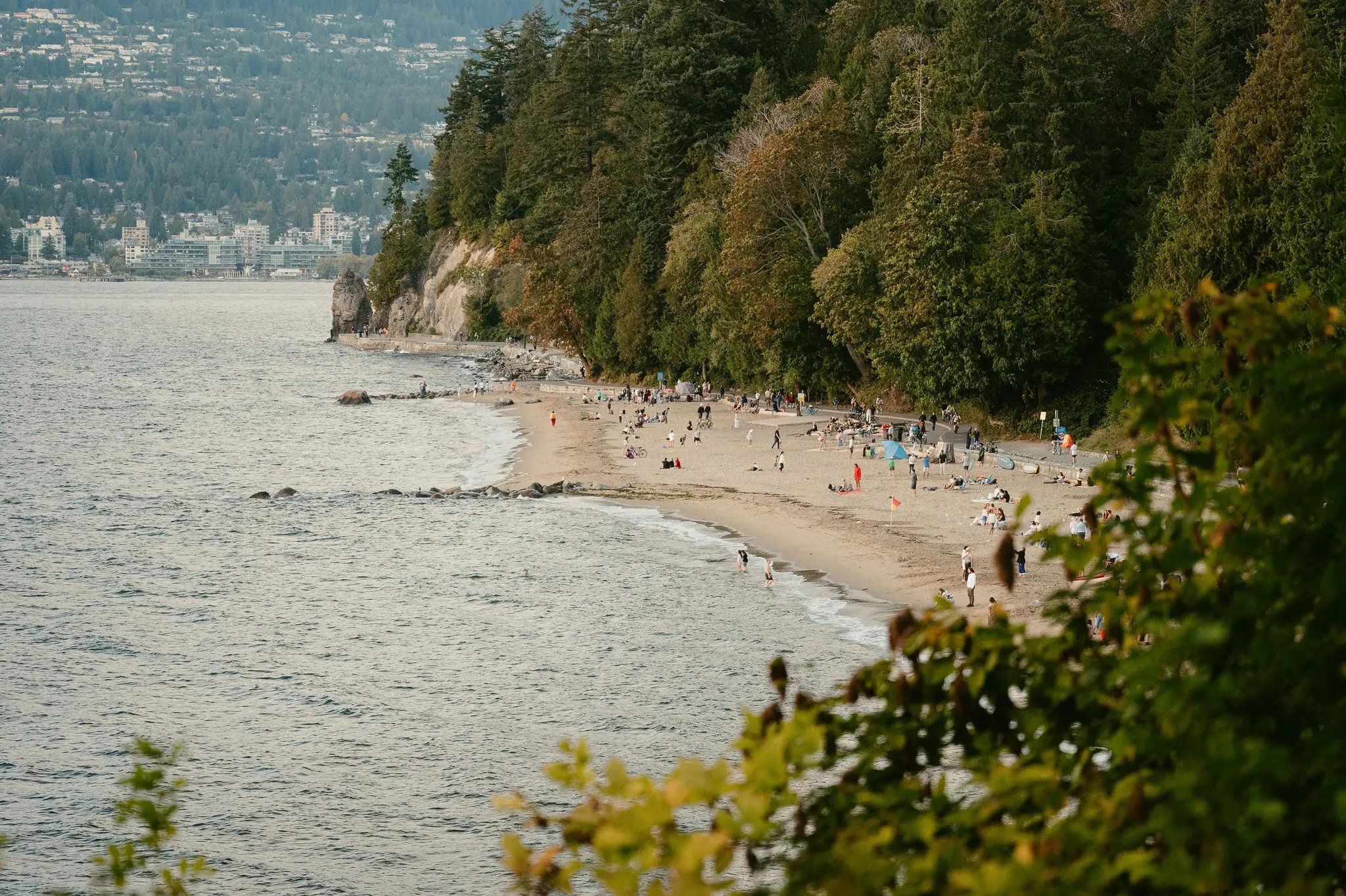 Sandy beach with people walking and relaxing near a forested area and calm water, city buildings visible in the background.