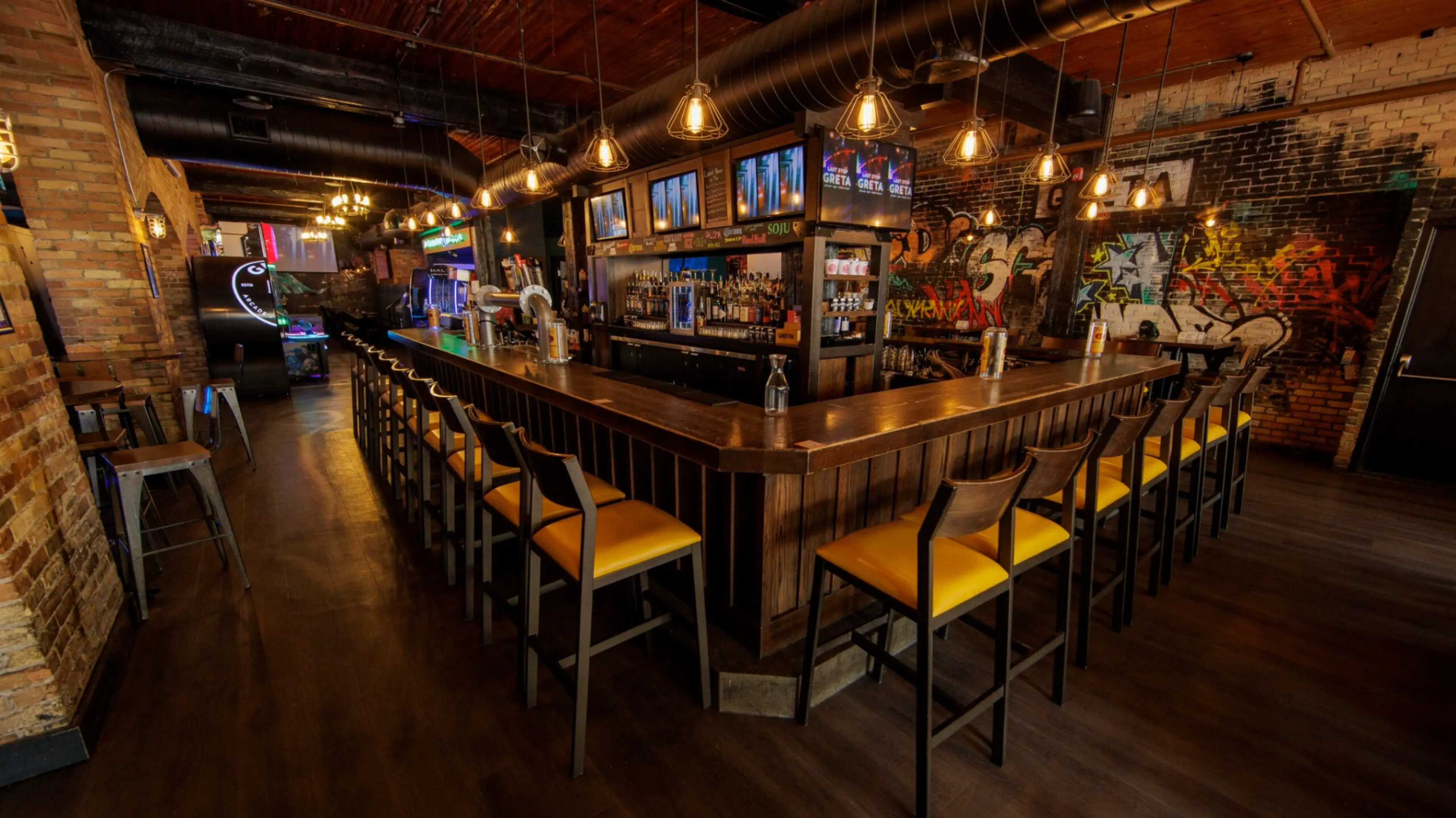 Empty bar with wooden counter, yellow cushioned stools, hanging lights, and graffiti on brick walls.