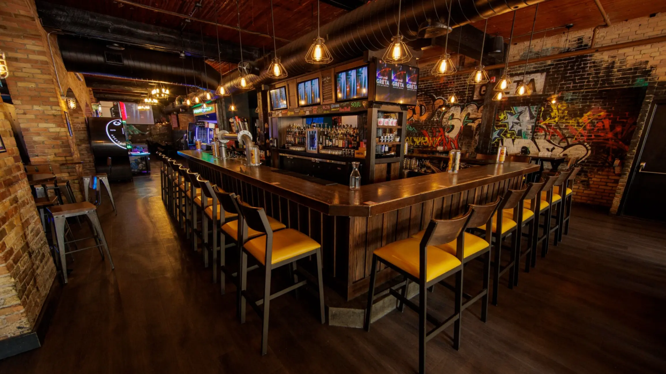 Empty bar with wooden counter, yellow cushioned stools, hanging lights, and graffiti on brick walls.