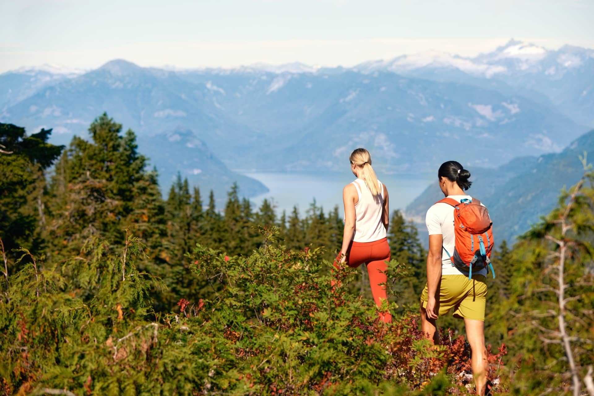 Two hikers walking through dense green forest with mountains and a lake in the background.